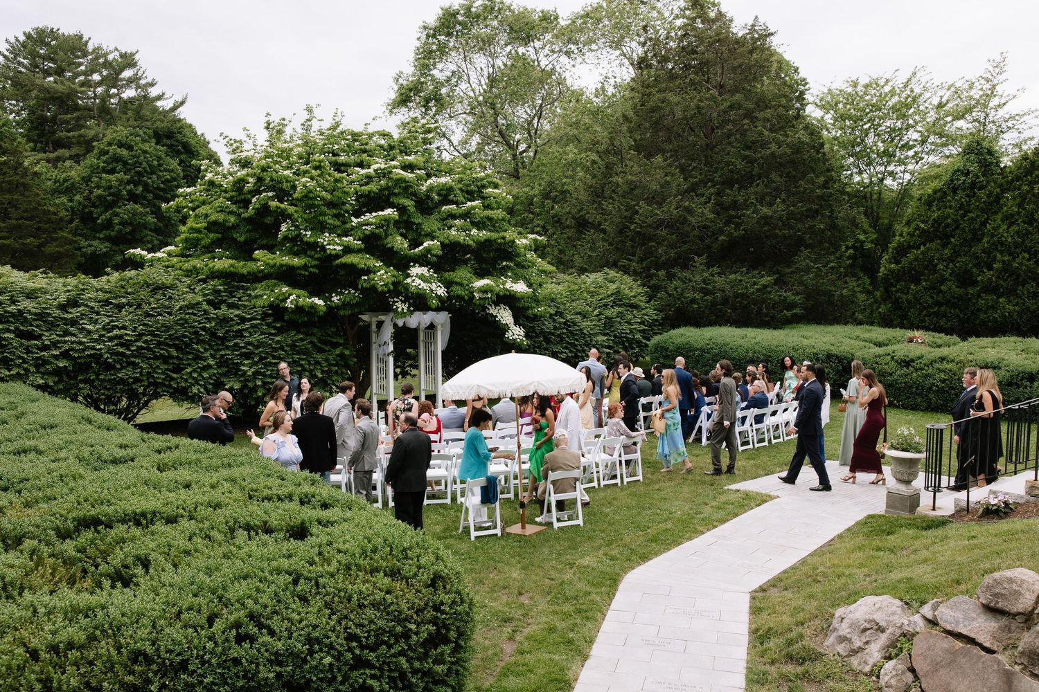 Outdoor wedding ceremony in a lush garden with white chairs, guests gathered, and a floral arch on a cloudy day.