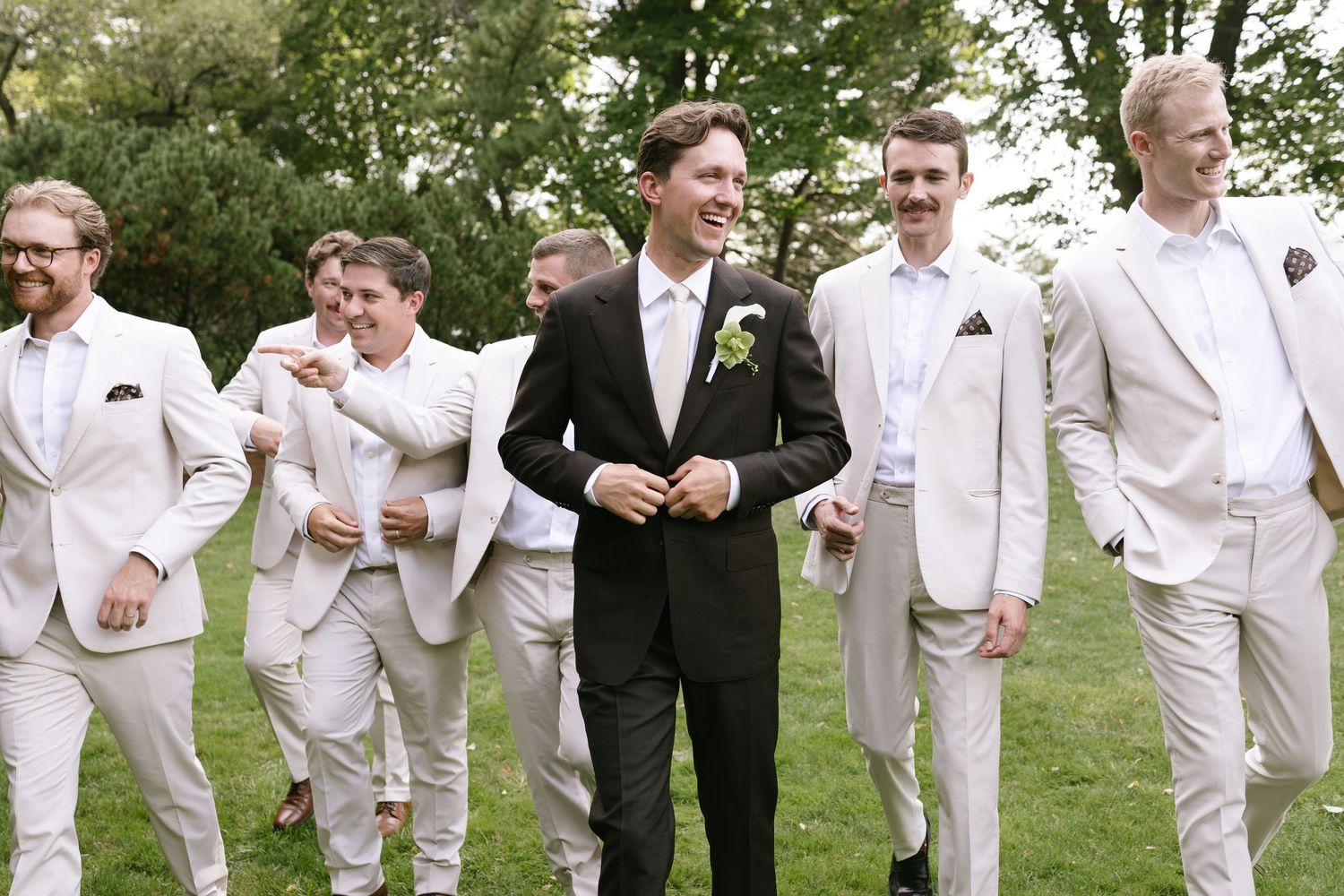 A smiling groom in a dark suit with a boutonniere walks across a lawn surrounded by groomsmen in light tan suits.
