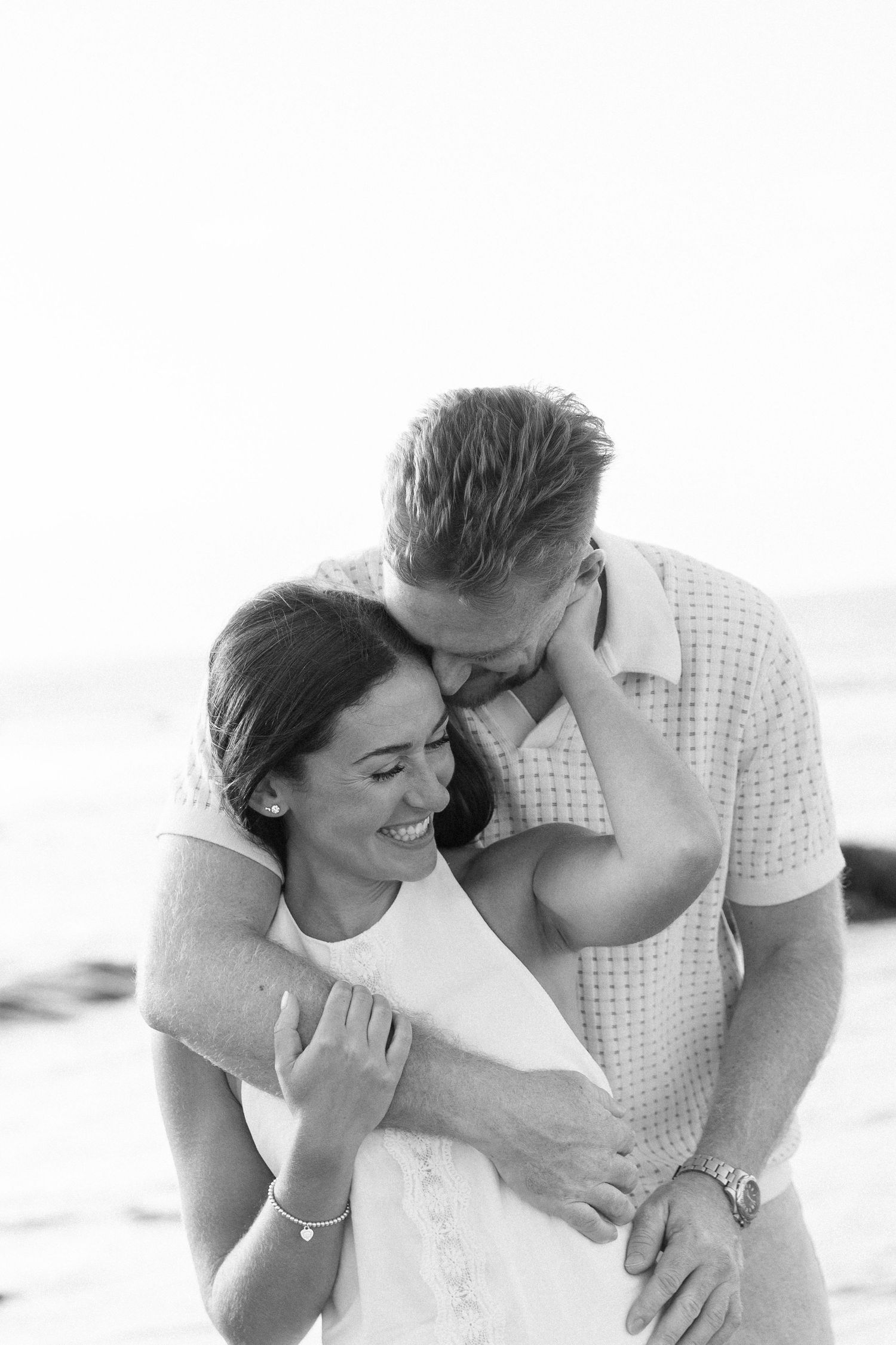 Black and white photo of a couple embracing on a beach, man kissing woman's temple as she laughs joyfully.