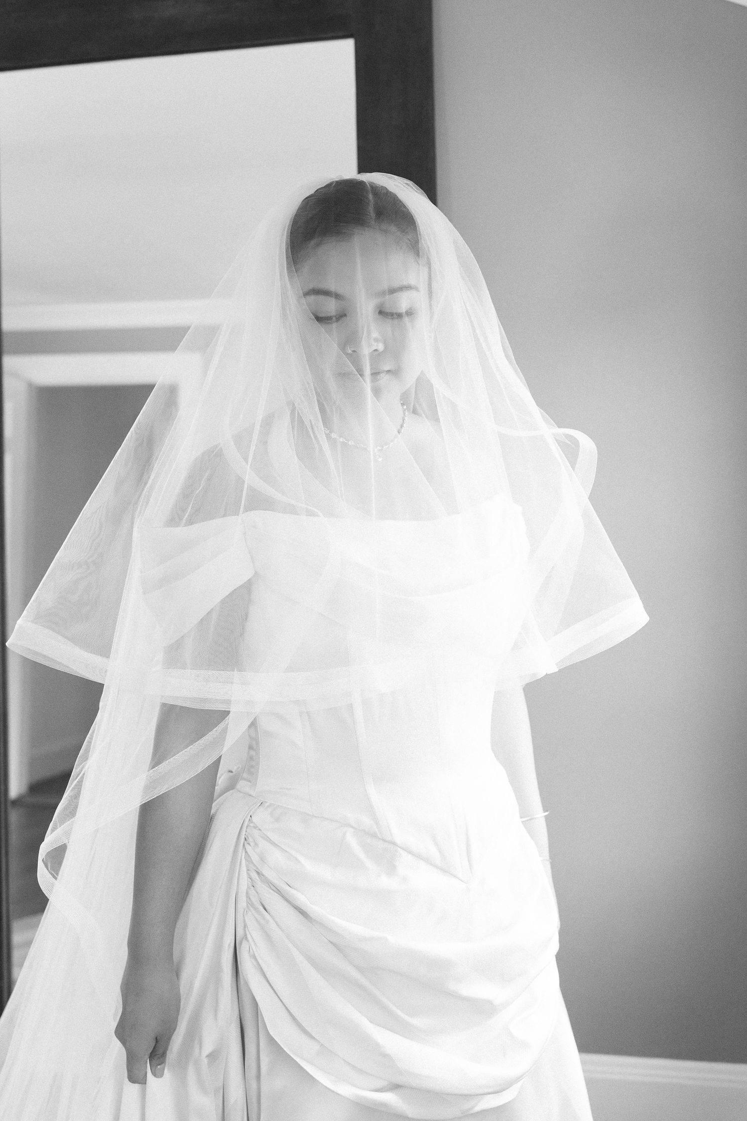 Black and white photo of a bride in a wedding dress and veil, eyes closed, standing in a softly lit room.