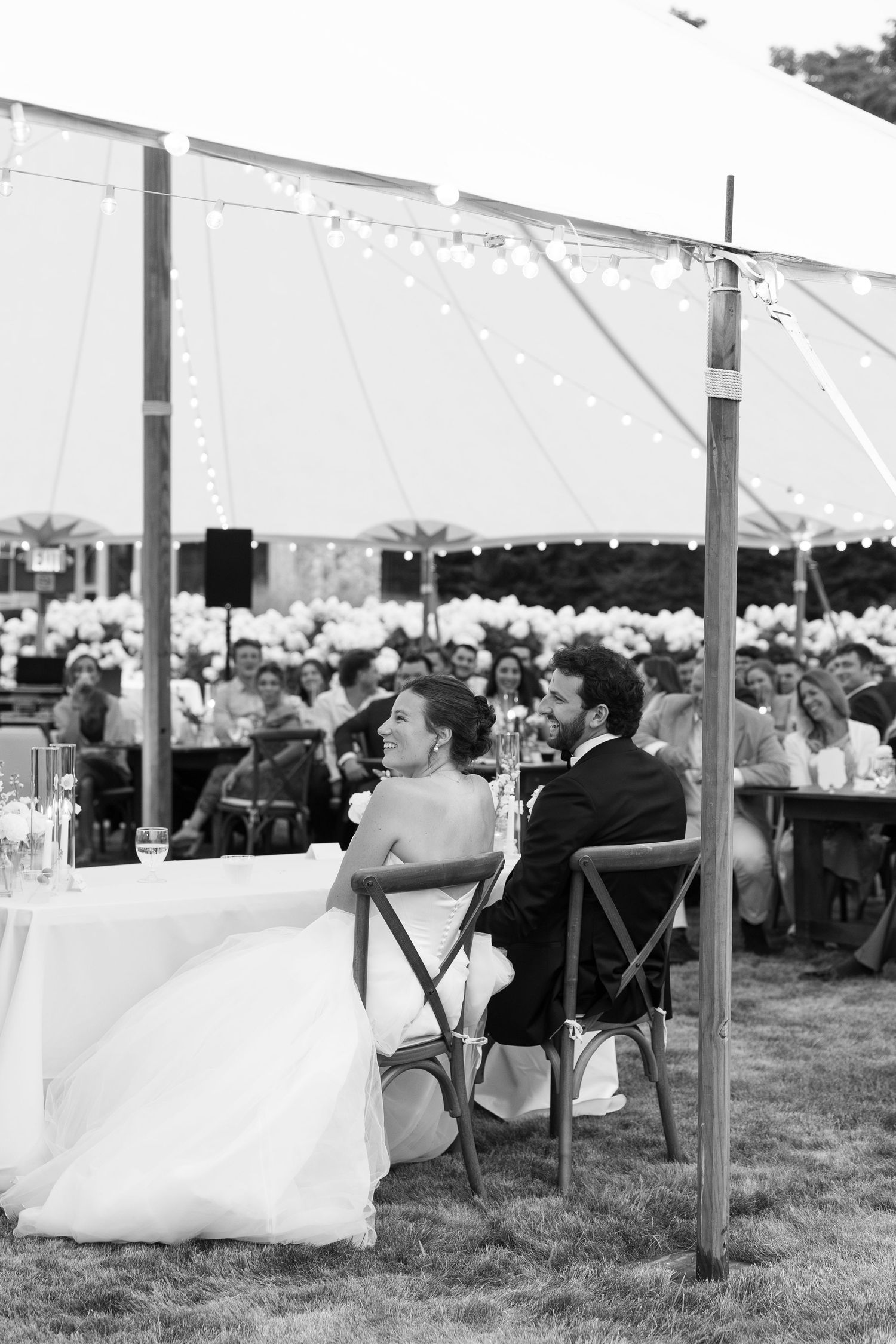 Black and white photo of bride and groom seated at sweetheart table during outdoor tented wedding reception.