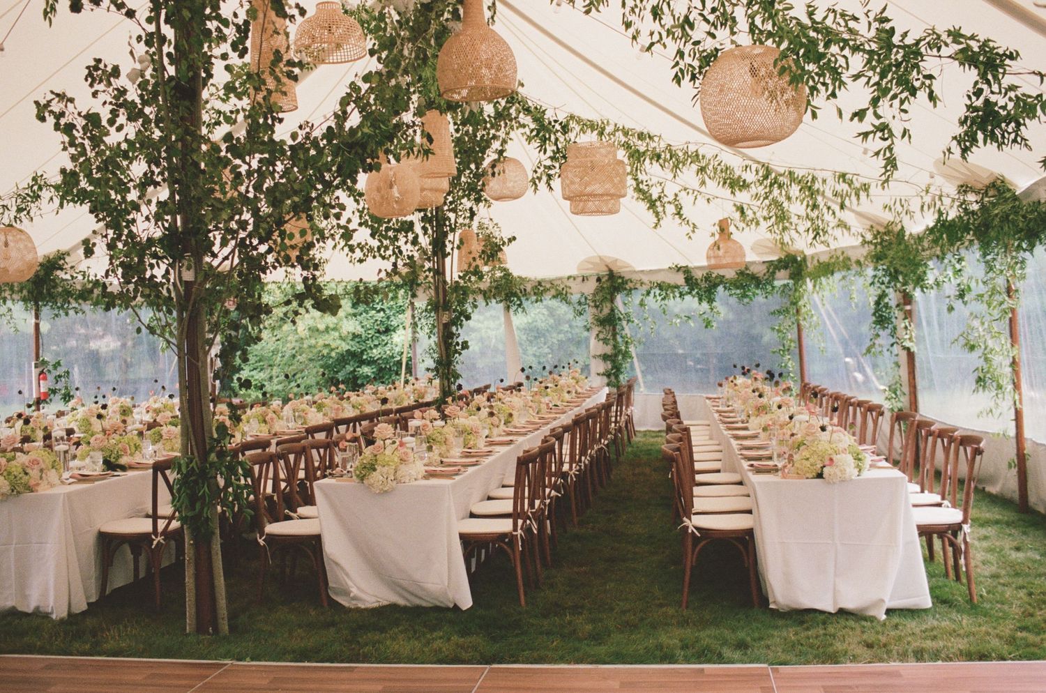 Elegant outdoor wedding reception under a white tent with long tables, lush greenery, wicker lanterns, and floral centerpieces.