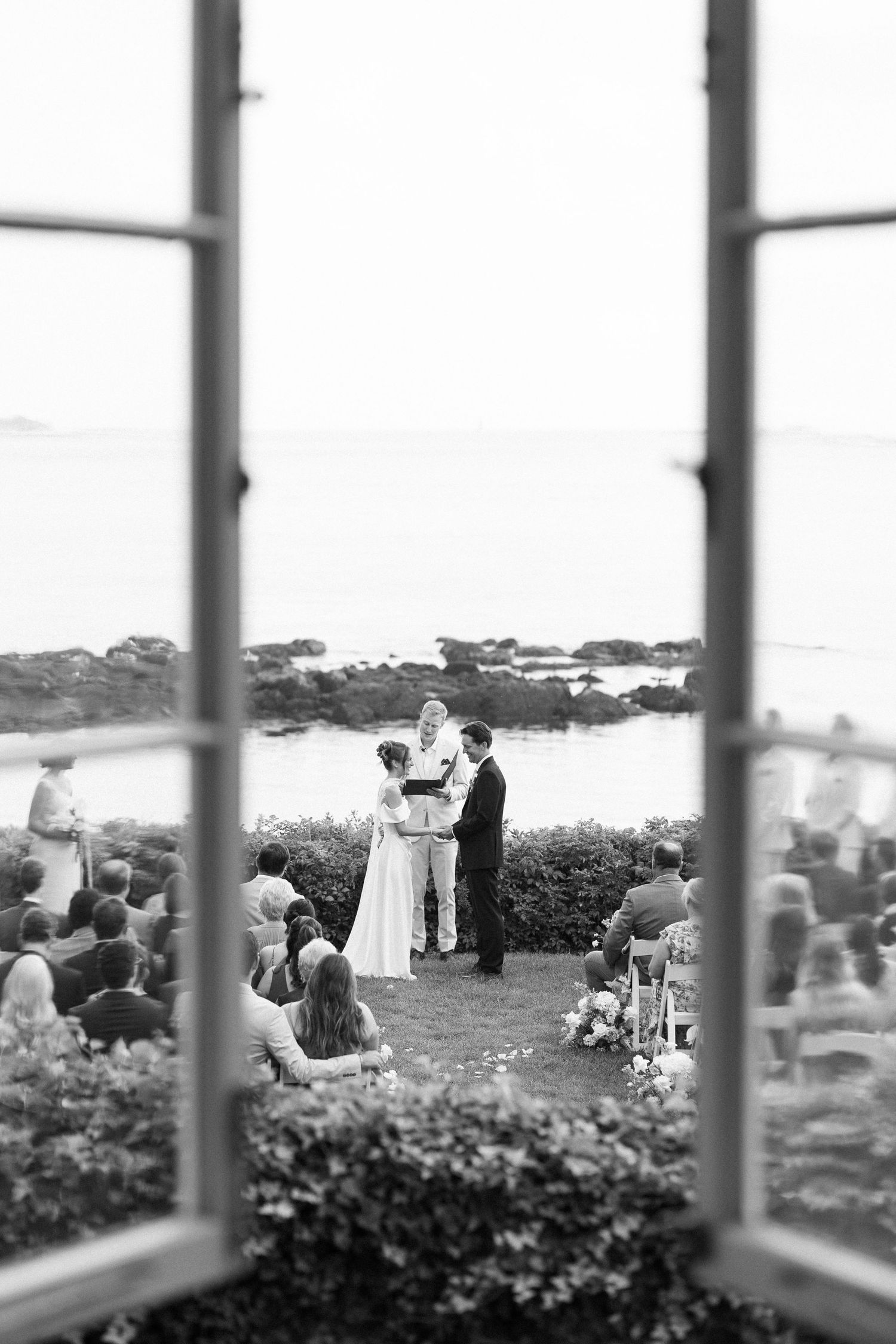 Black and white photo of outdoor waterfront wedding ceremony viewed through open window.