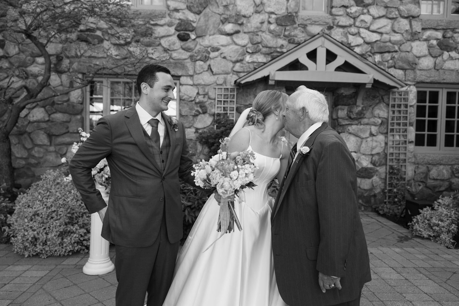 A black and white wedding photograph showing a couple and an officiant outside a stone building with rustic details.
