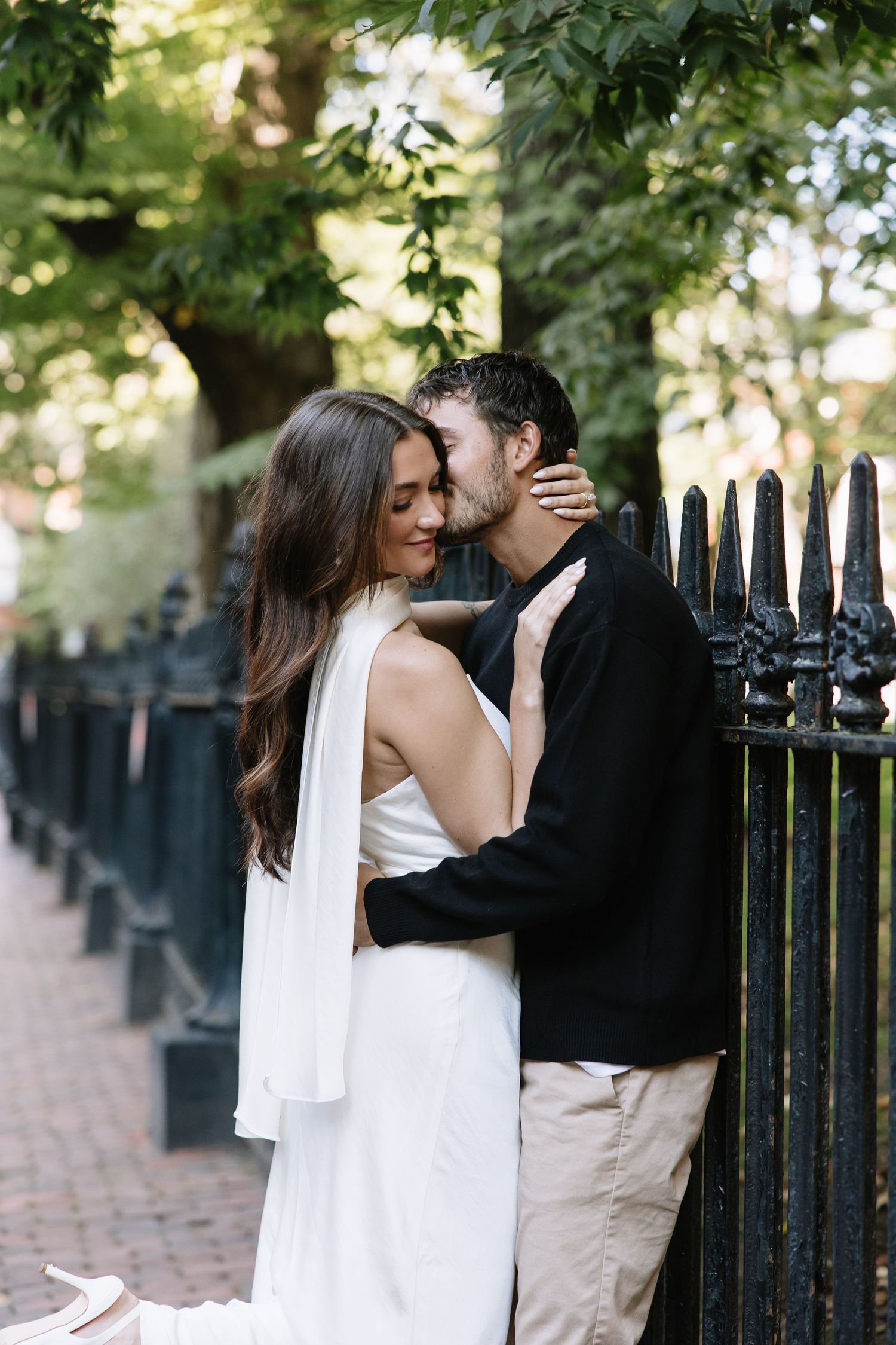 Romantic couple embracing by a black iron fence in an outdoor garden setting.