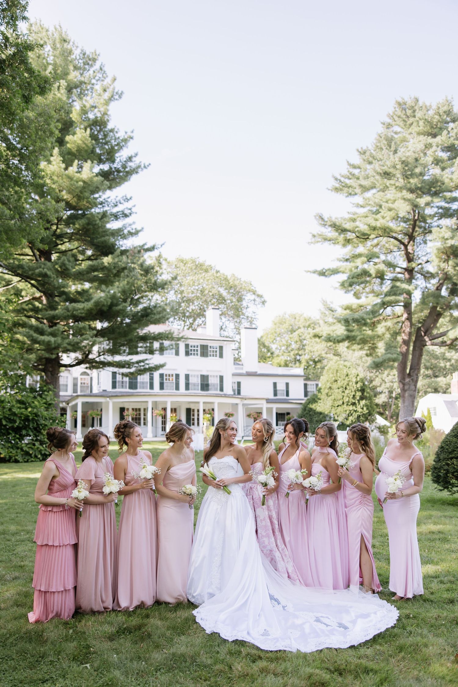 Wedding party in pink gowns gathered on lawn in front of classic white colonial mansion.