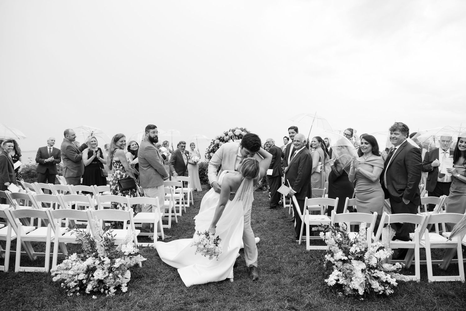 Wedding guests seated in white chairs on a lawn for an outdoor ceremony.