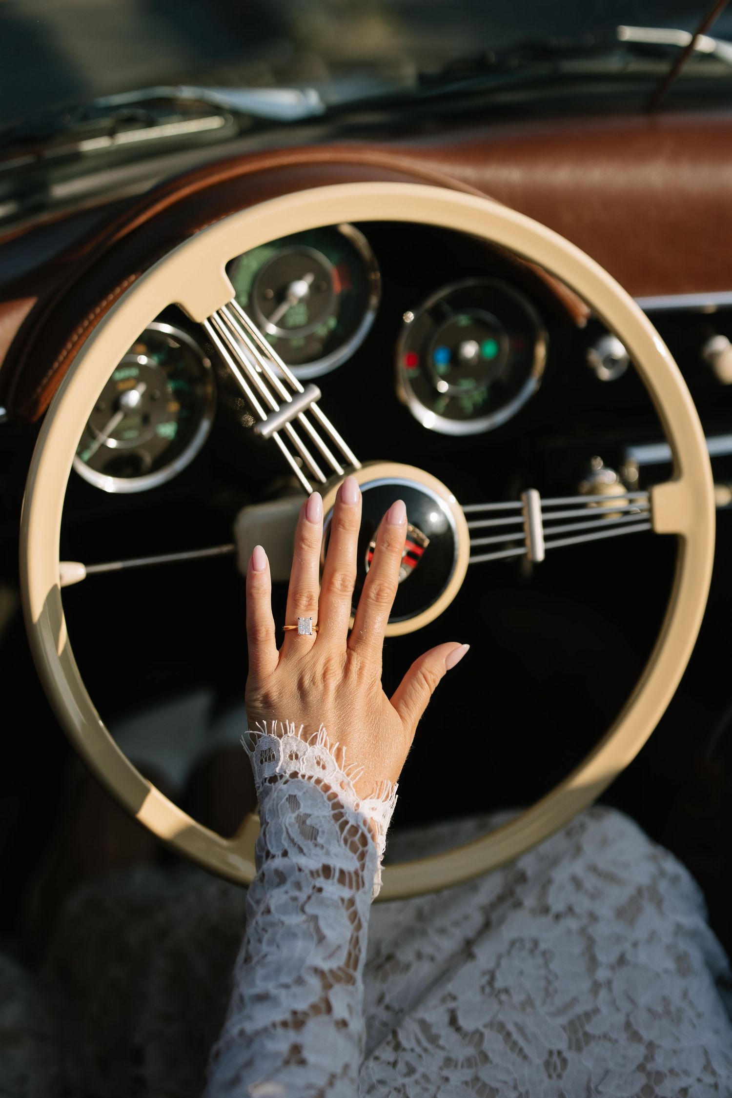 Bride's hand with ring on vintage car steering wheel, wearing lace wedding dress sleeve in classic retro vehicle interior.
