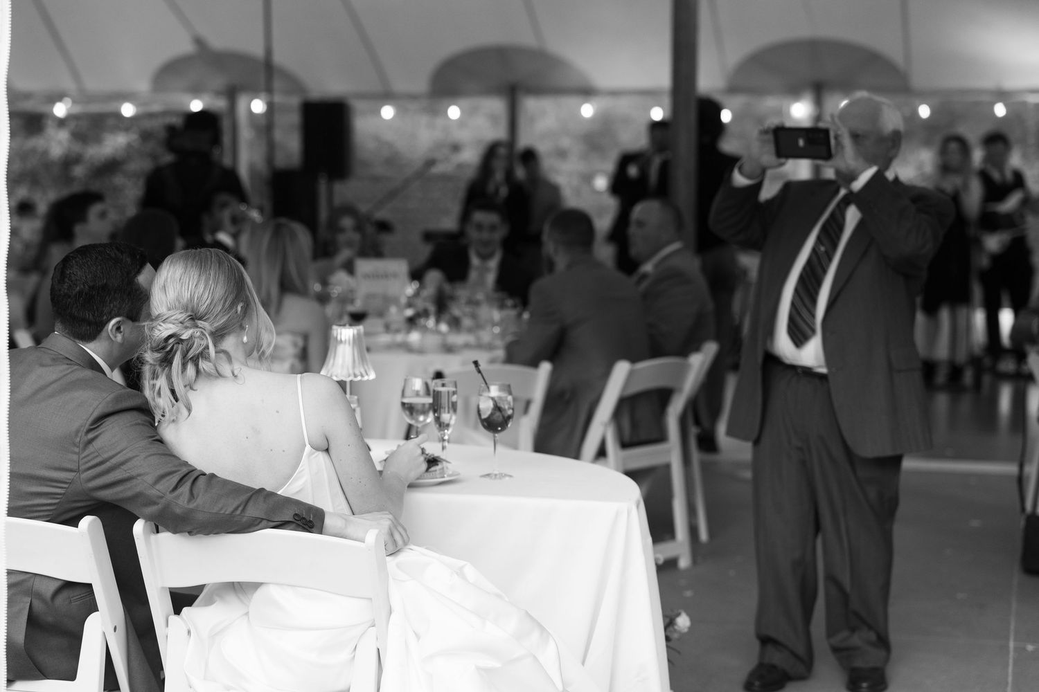 Black and white wedding reception photo with guests seated at tables while a man in a suit takes a photo.
