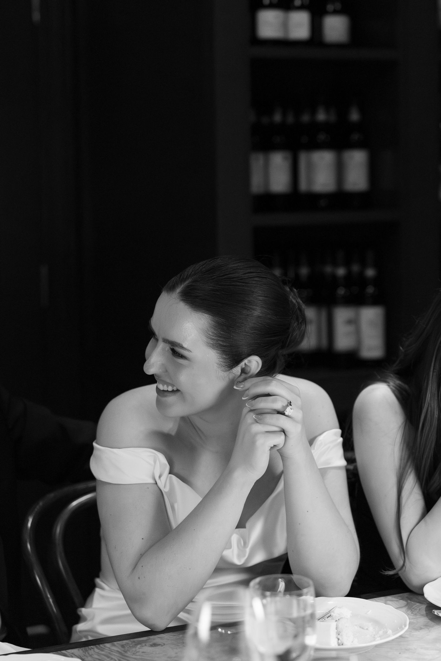 Black and white photo of a smiling woman in off-shoulder dress seated at a restaurant table with wine bottles in background.