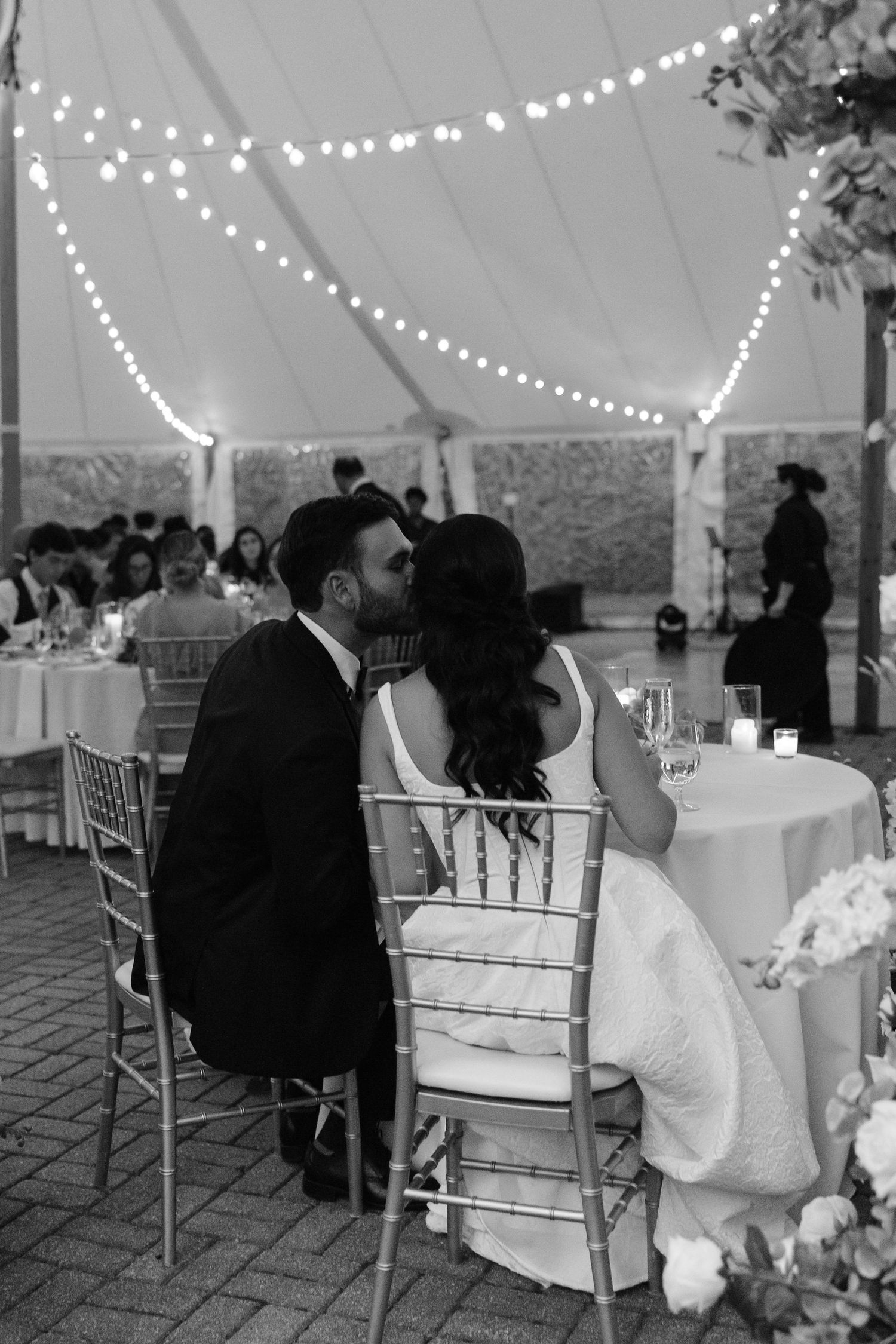 Black and white photo of a bride and groom sharing a kiss at their wedding reception under string lights.