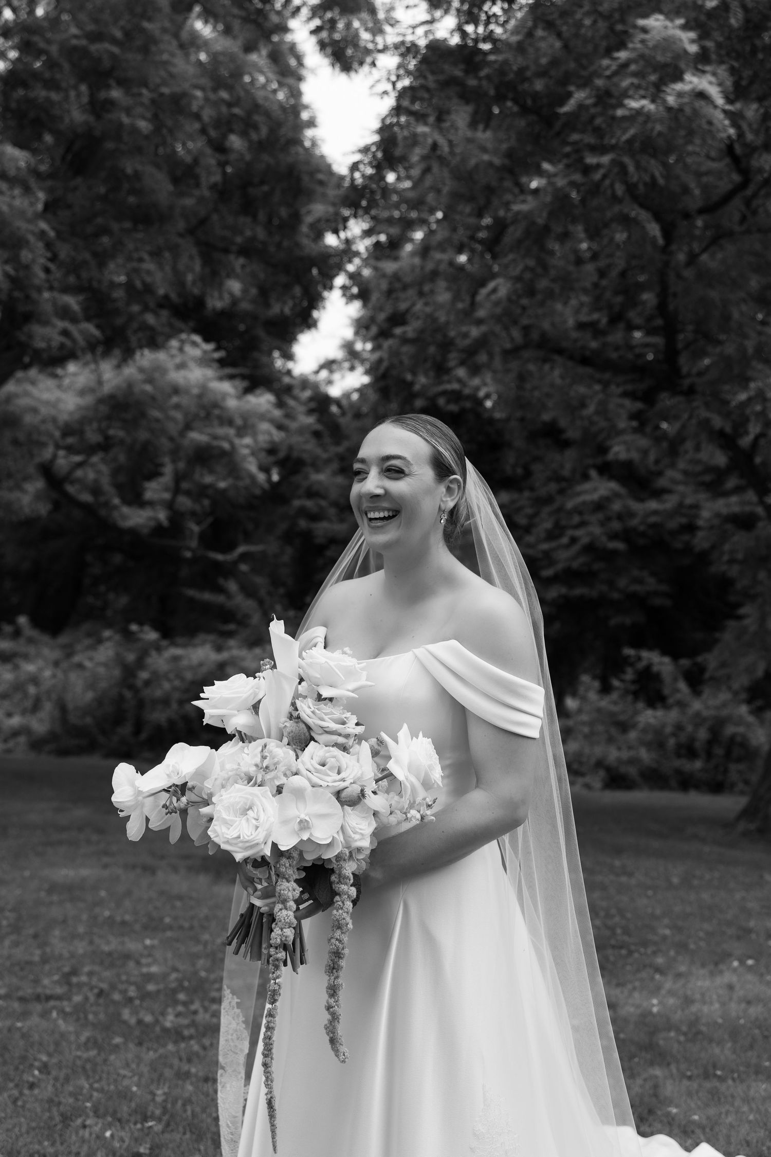 Joyful bride in off-shoulder gown and veil holding white floral bouquet outdoors in black and white photo.
