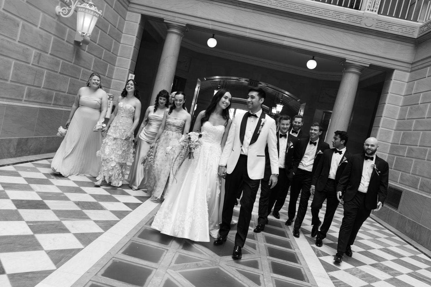 Black and white wedding party photo with bride, groom, bridesmaids, and groomsmen walking in elegant venue.