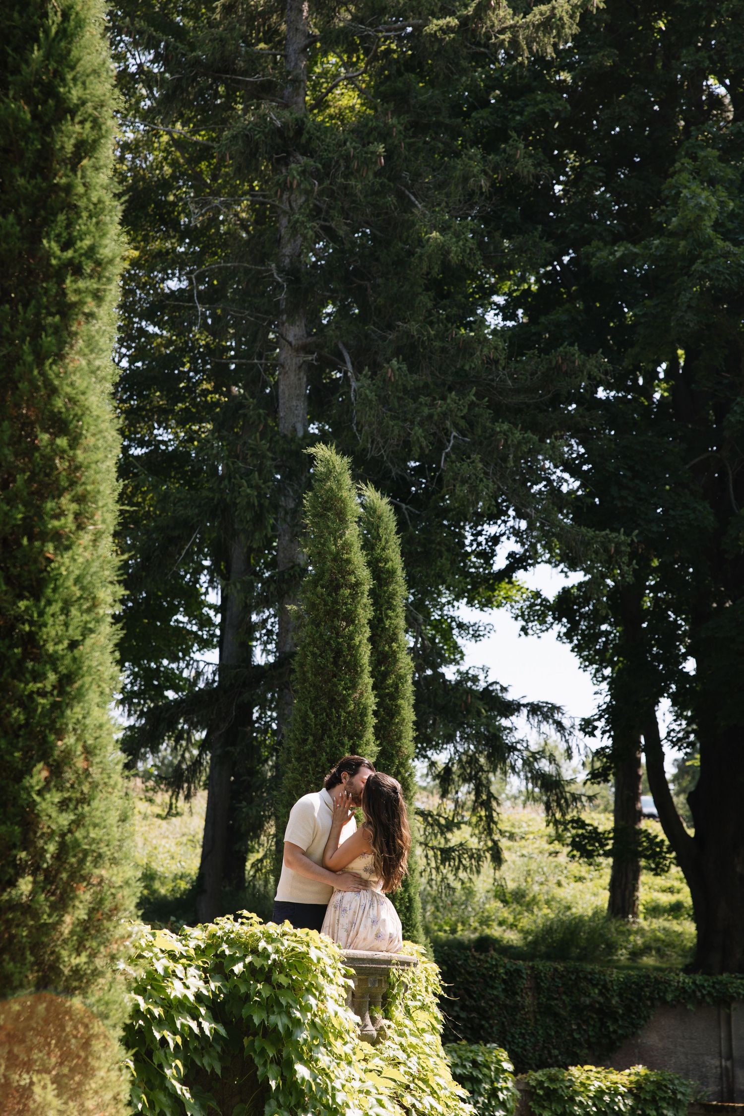 Couple sharing a romantic kiss surrounded by tall cypress and pine trees in a lush green garden setting.