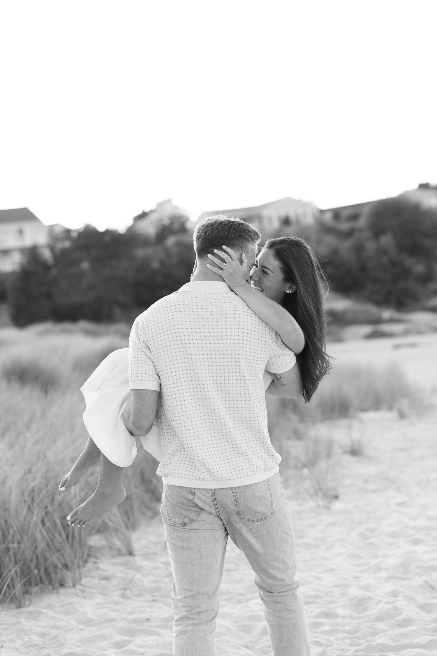 Black and white photo of a couple embracing and kissing on a sandy beach with dunes and trees in the background.
