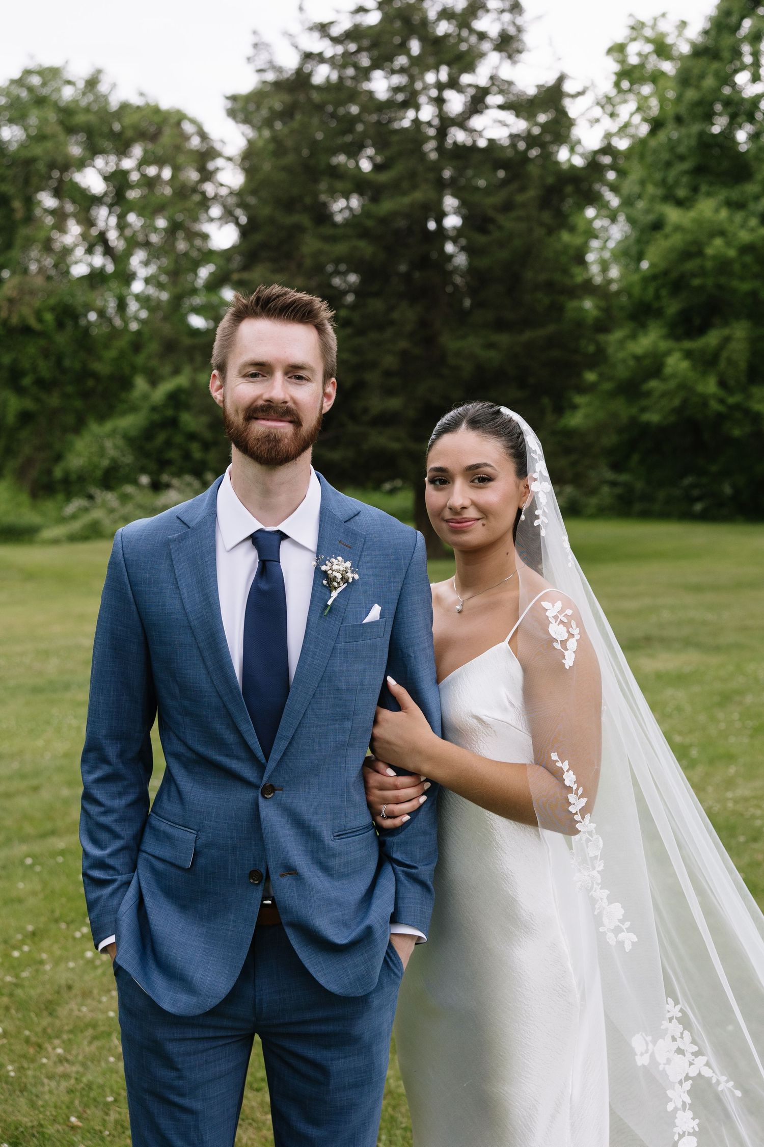 A bride and groom pose outdoors on green grass, she in a white gown with veil, he in a blue suit with navy tie.