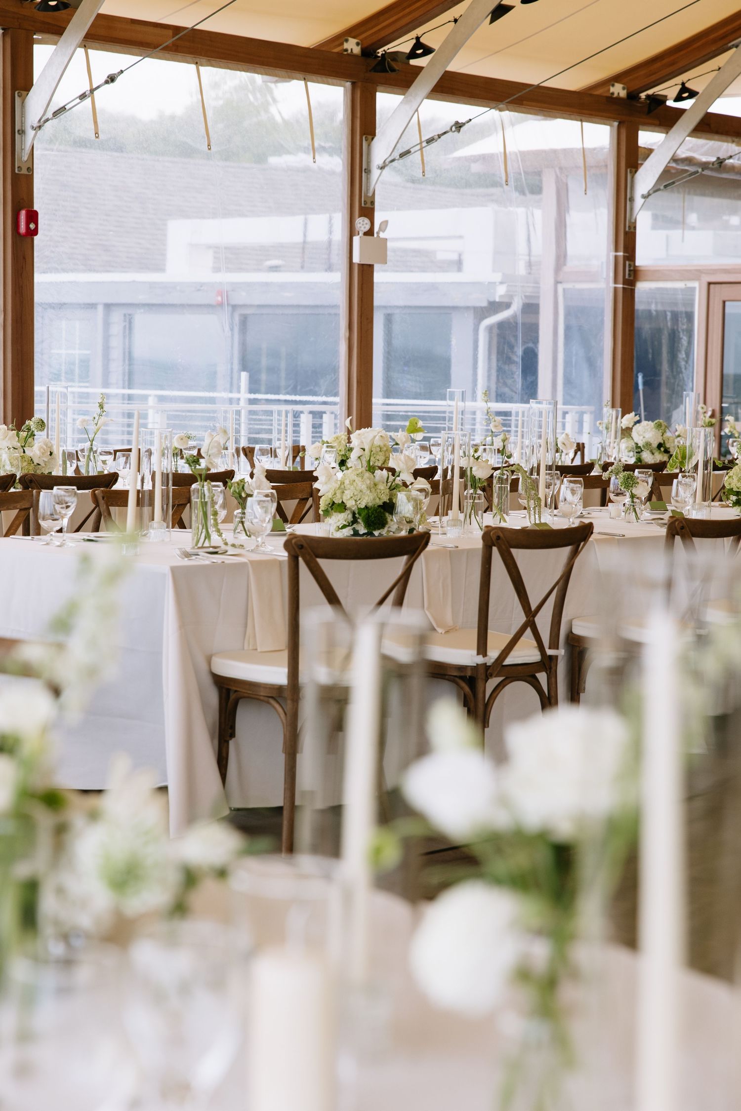 Elegant wedding reception with white linen tables, wooden cross-back chairs, and white floral centerpieces in a bright glass venue.