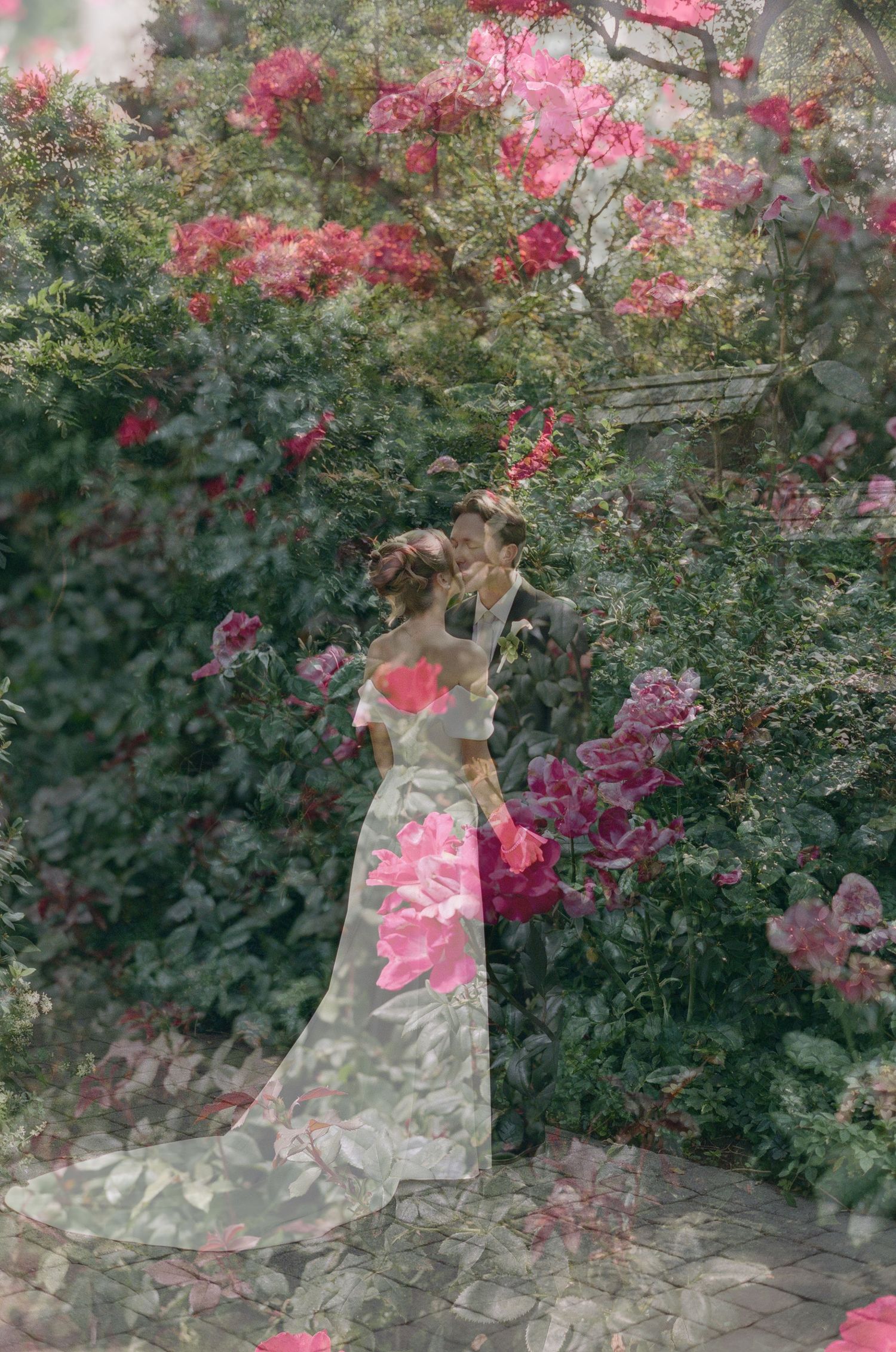 Double exposure wedding photo of a bride in a long white gown embracing a groom, blended with pink flowers and green garden foliage.