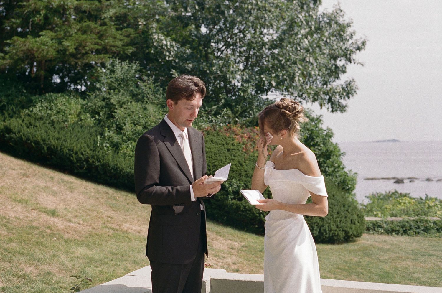 A groom in a dark suit and a bride in a white gown read private letters outdoors on a lawn overlooking the water.