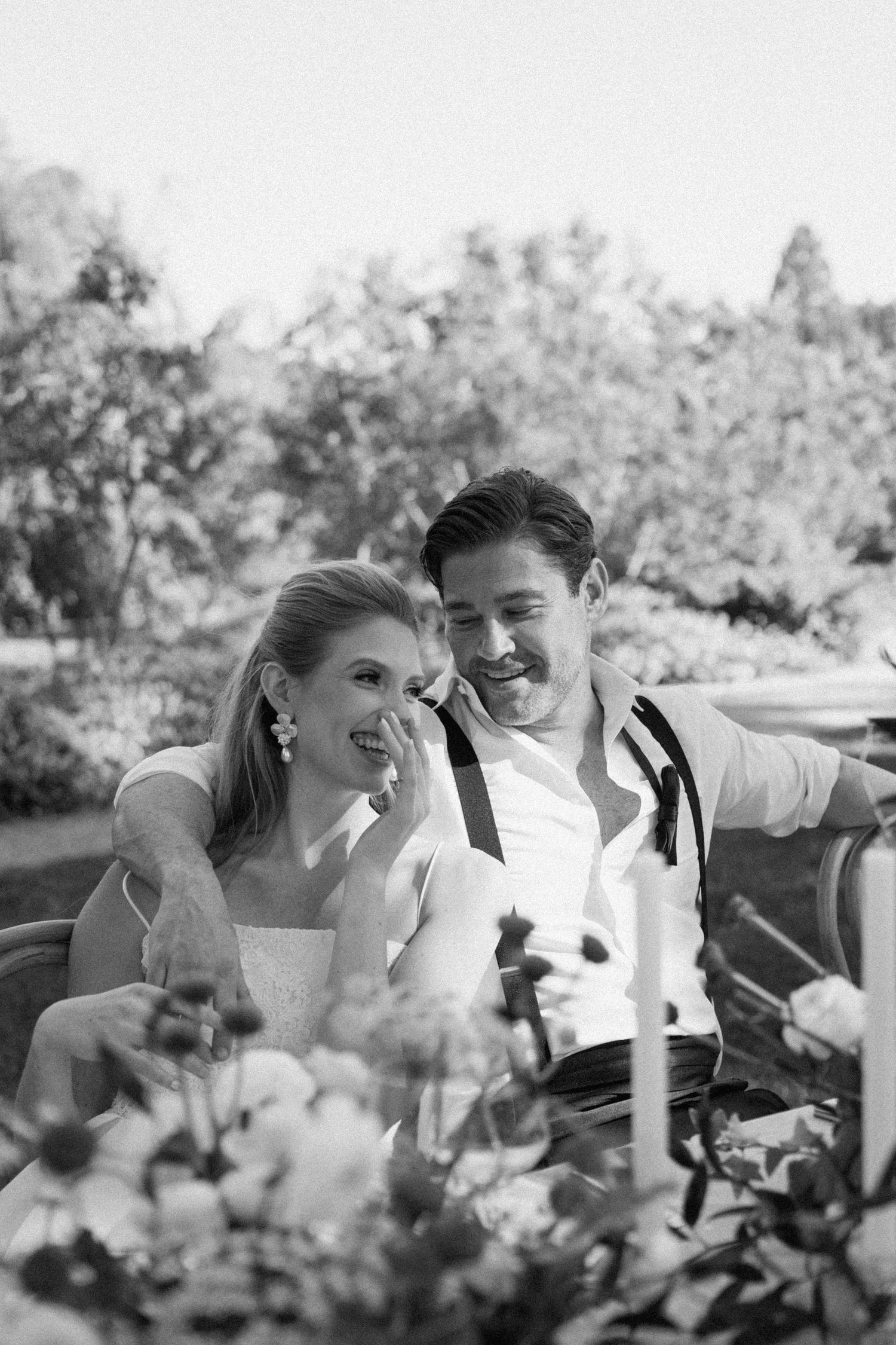 Black and white photo of a smiling bride and groom sitting together at a floral wedding reception table outdoors.