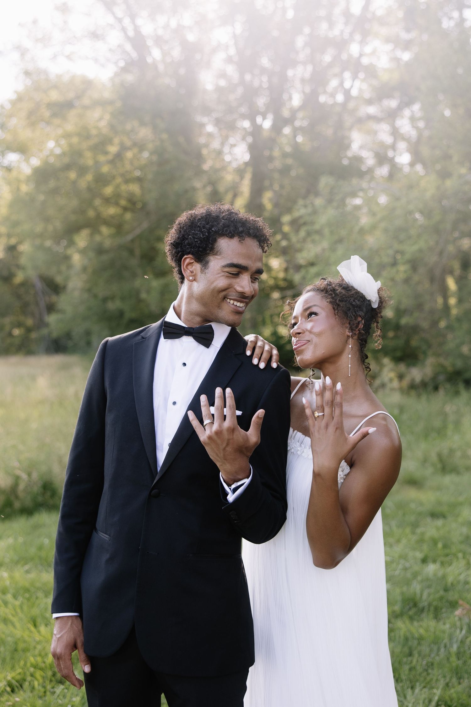 Happy newlyweds in formal attire show off wedding rings outdoors, groom in tuxedo and bride in white dress.