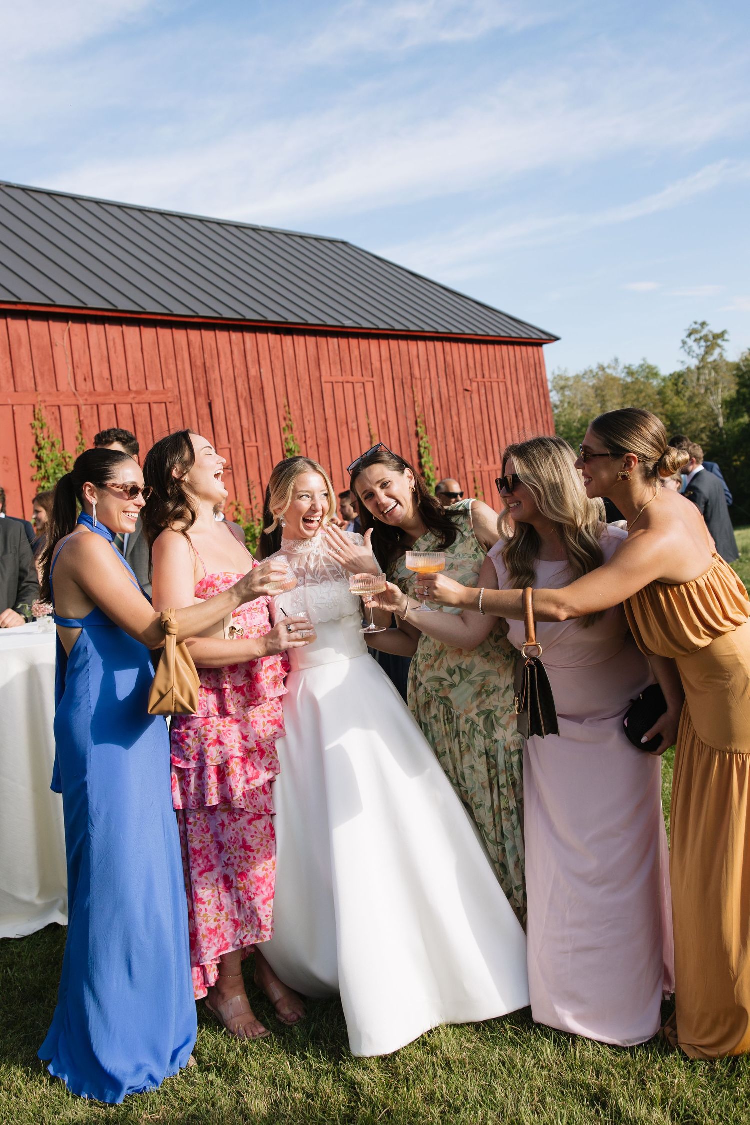 A bride in a white gown laughs joyfully with bridesmaids in colorful dresses outside a red barn.