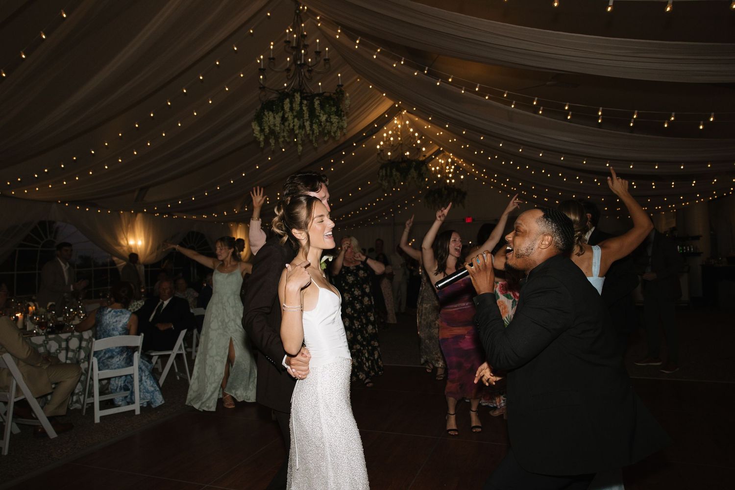 Bride and groom dancing joyfully with guests under twinkling fairy lights at an elegant wedding reception.