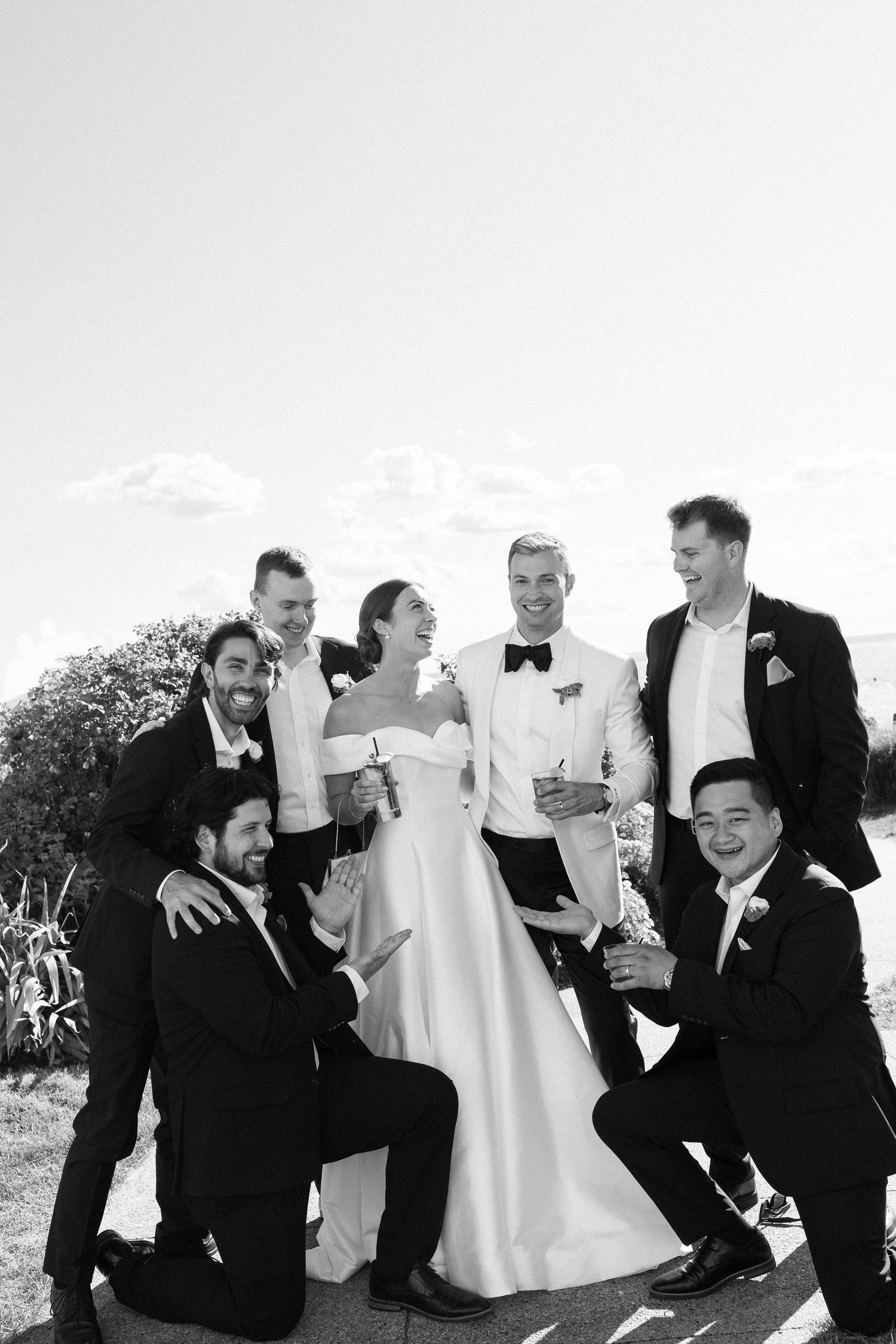 Black and white wedding photo of bride and groom with groomsmen posing outdoors, some kneeling, all smiling joyfully.