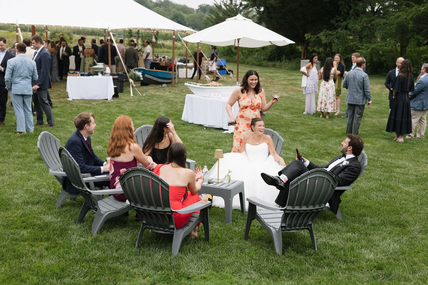 Bride and wedding guests relaxing in Adirondack chairs on grass at outdoor wedding cocktail hour.