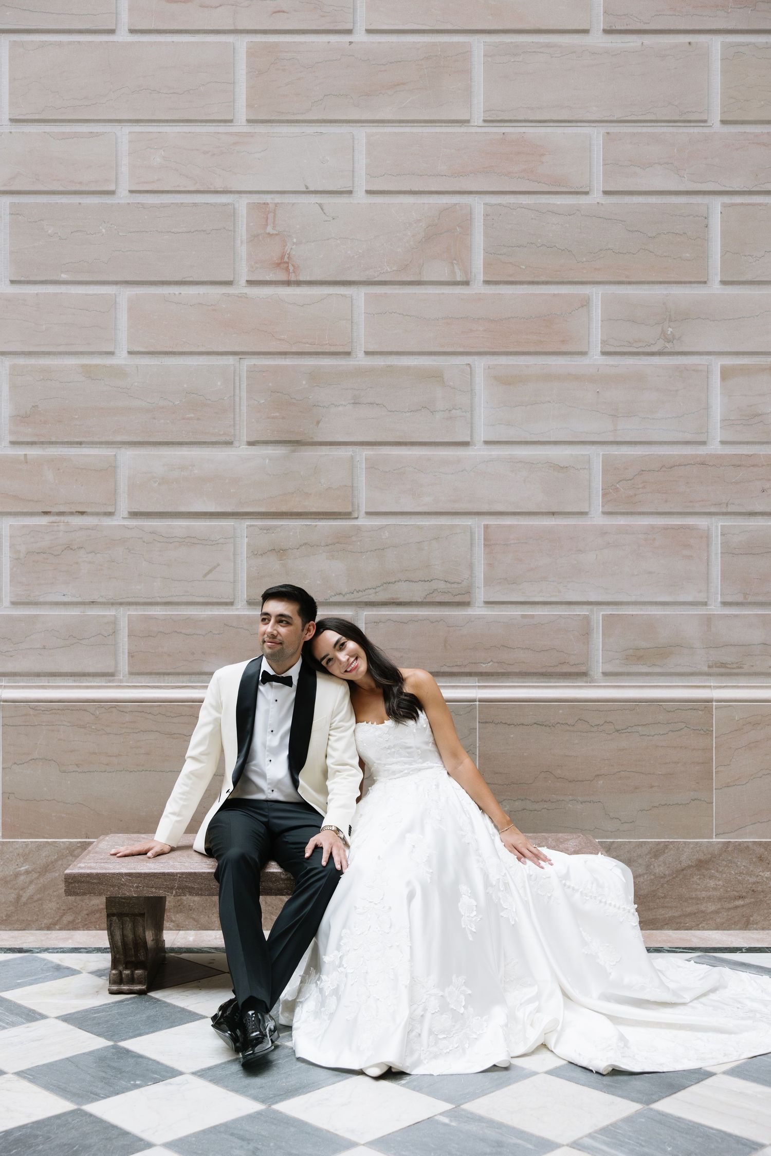 Bride and groom sitting on a bench against a stone wall, bride leaning on groom in elegant wedding attire.