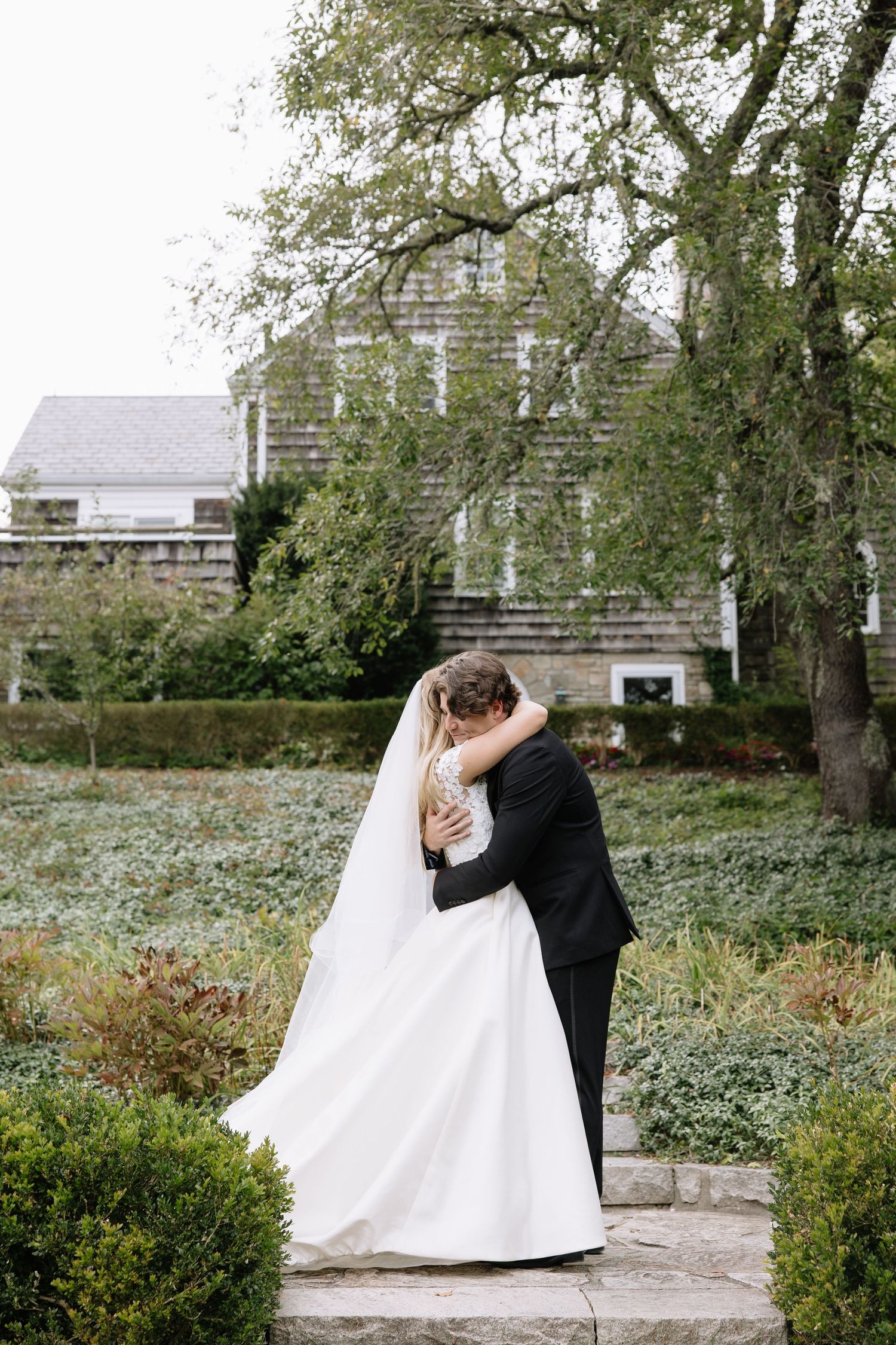 A romantic wedding portrait of a couple embracing in front of a rustic barn with overhanging trees.