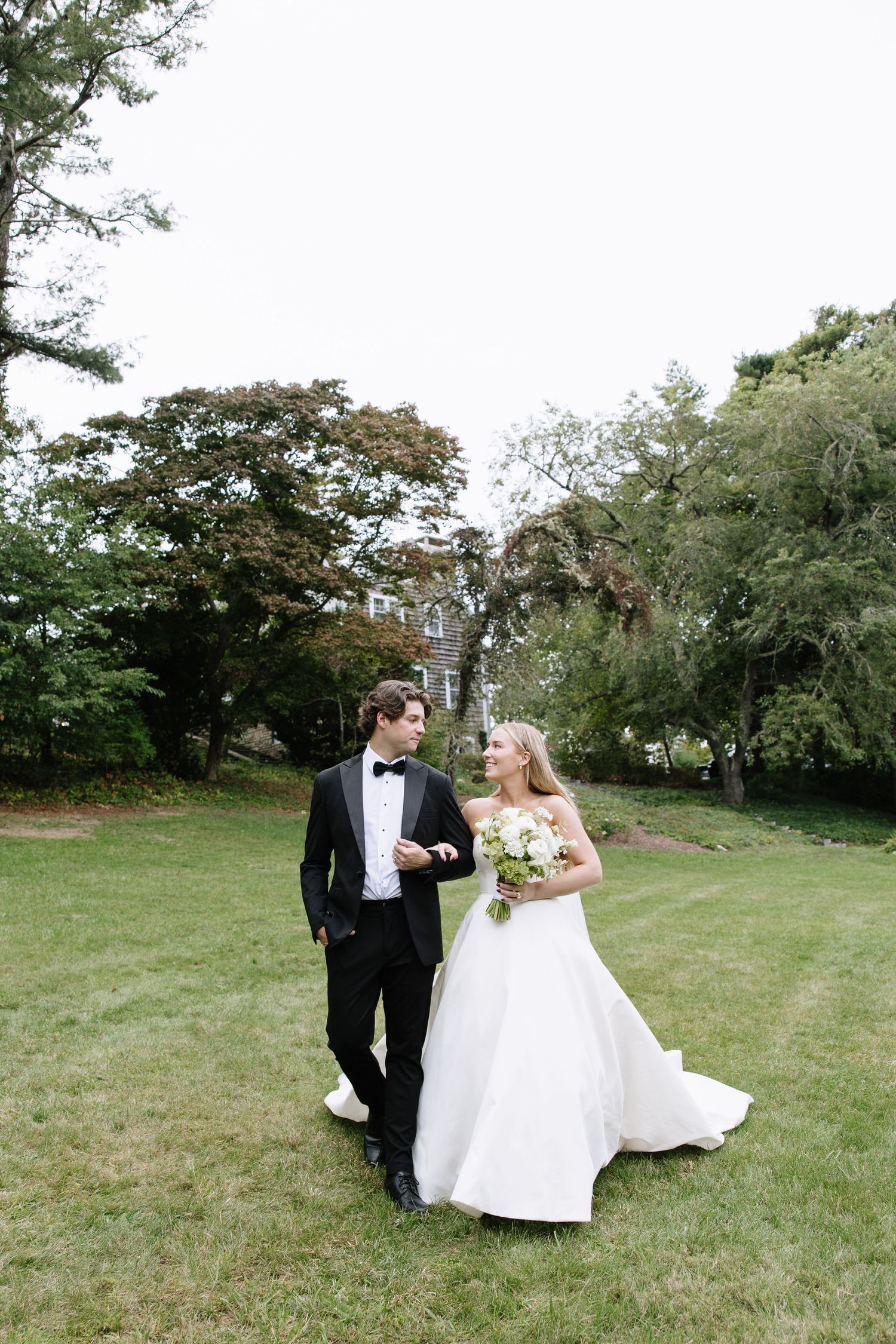 Wedding couple poses together on a grassy lawn surrounded by trees.