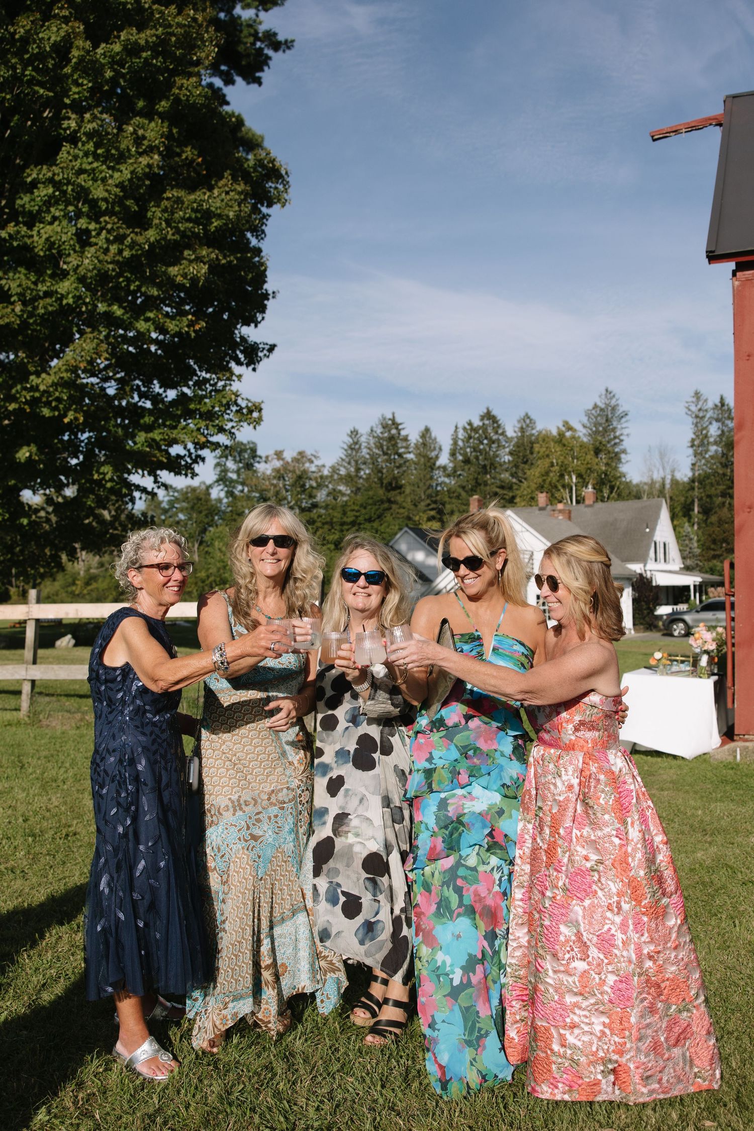Five women in elegant dresses toasting with champagne glasses outdoors at a sunny wedding celebration.