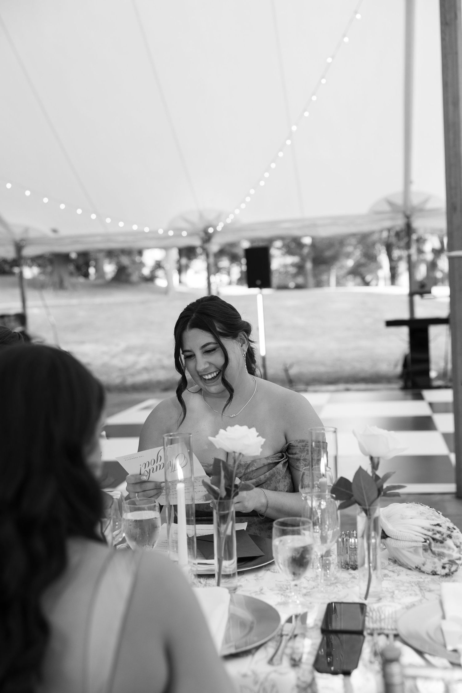 Black and white photo of a smiling bride holding a white flower at an elegant outdoor wedding reception table.