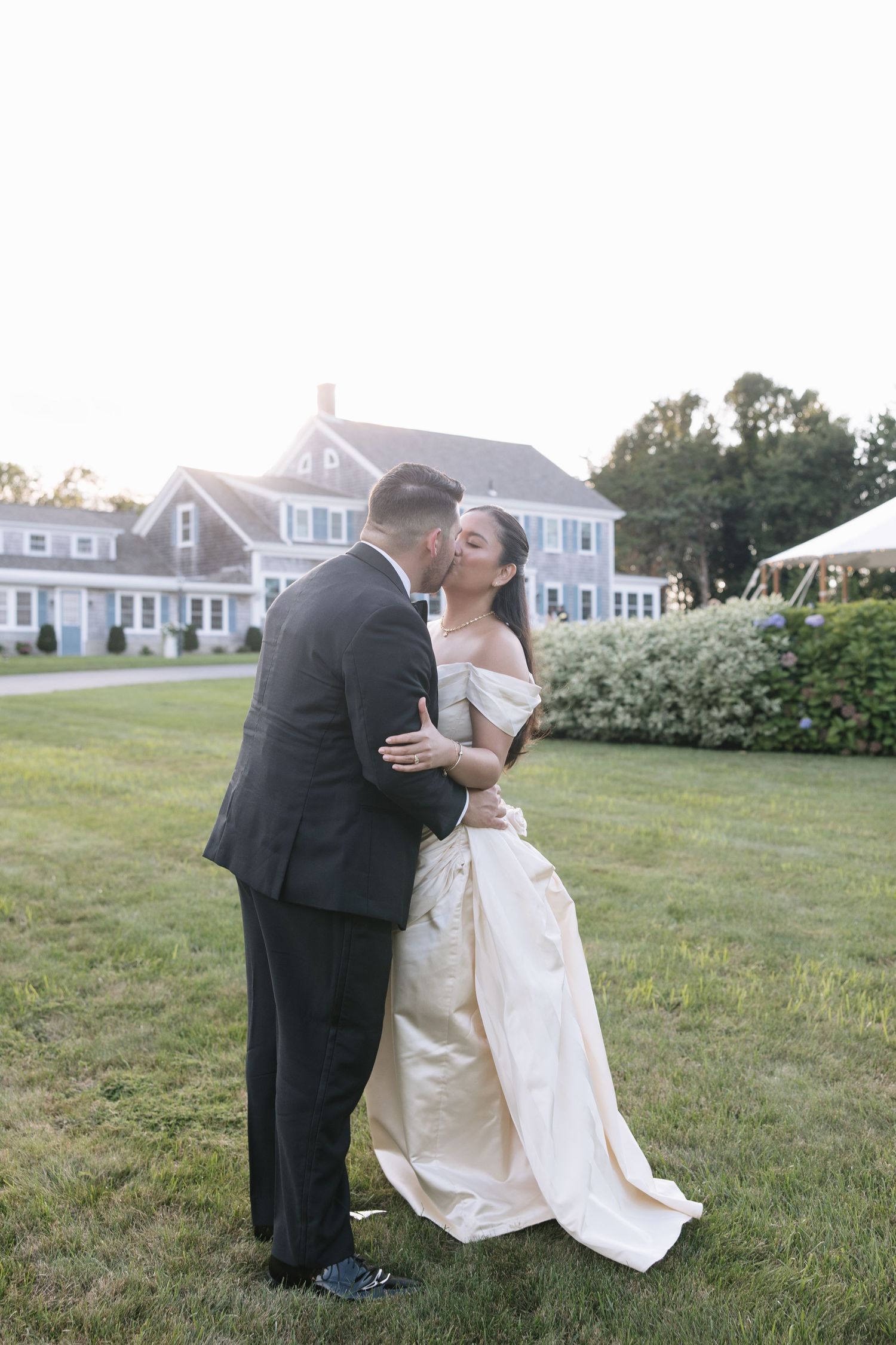 Bride and groom share a romantic kiss on green lawn outside elegant Cape Cod estate during golden hour wedding.
