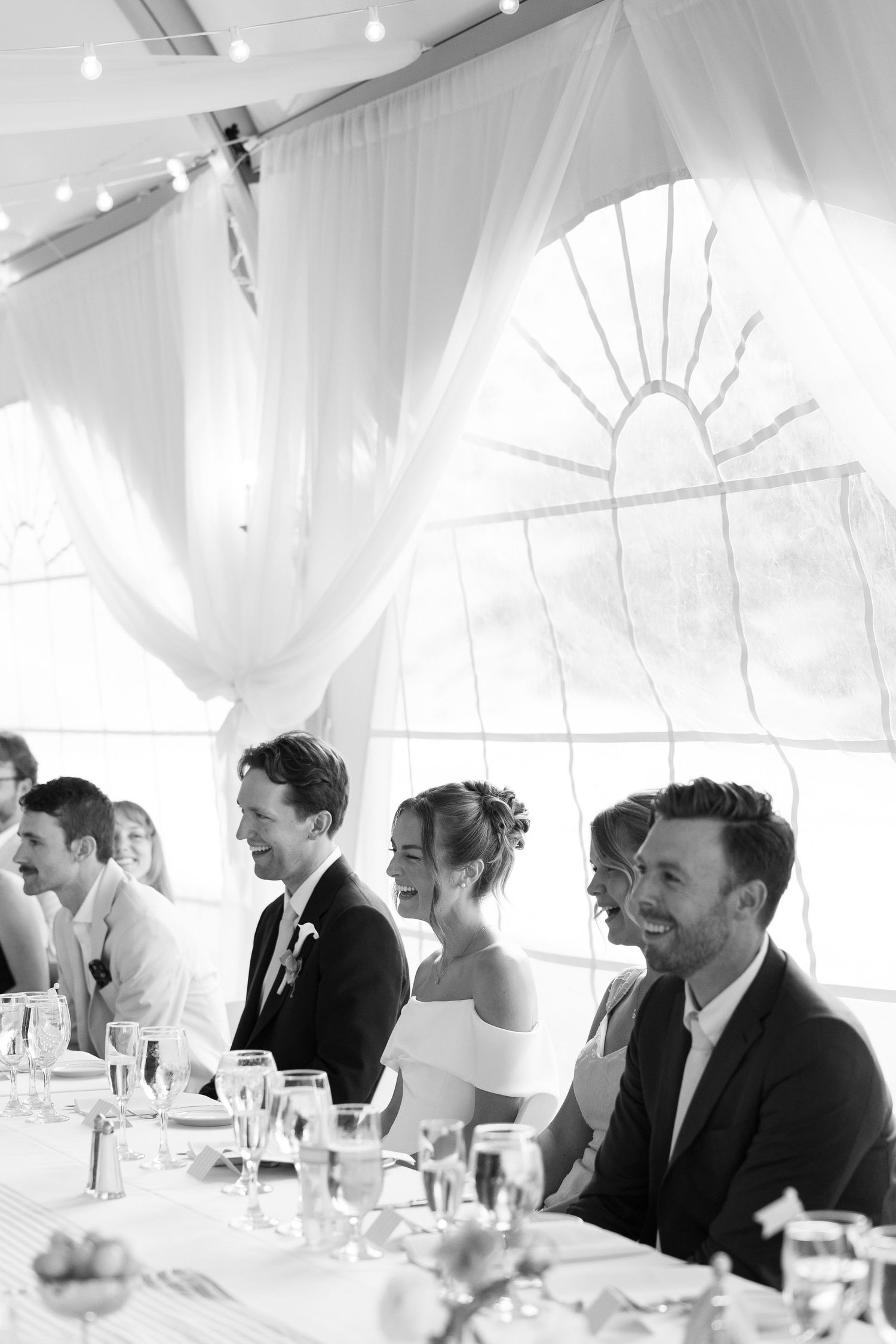 Black and white photo of smiling wedding party seated at reception table under elegant draped tent.