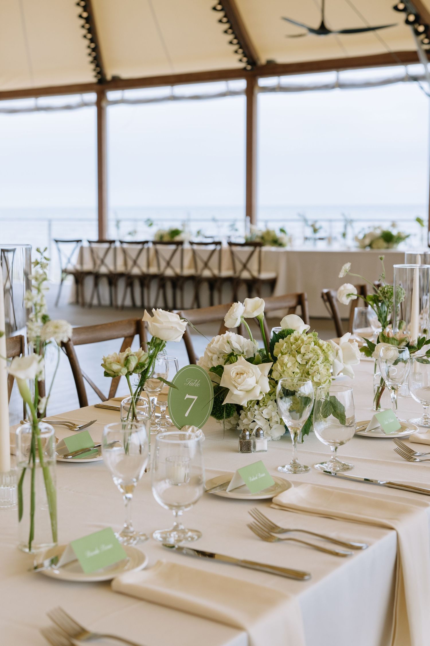 Elegant wedding reception table set with white roses, hydrangeas, green accents, and table number 7 overlooking the ocean.