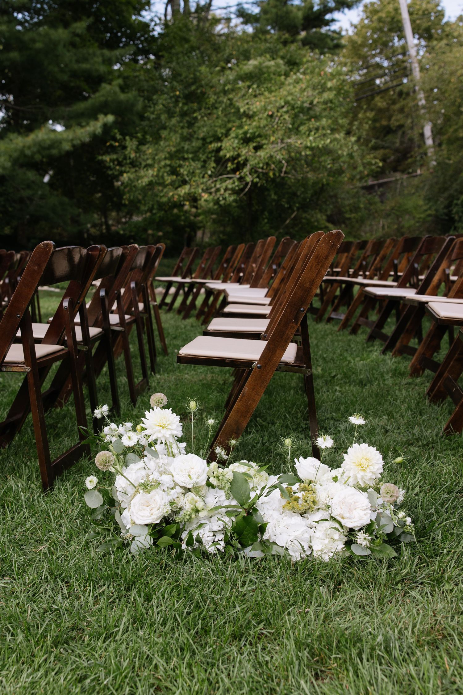 Wooden chairs arranged on green lawn with white floral aisle decorations for outdoor wedding ceremony.