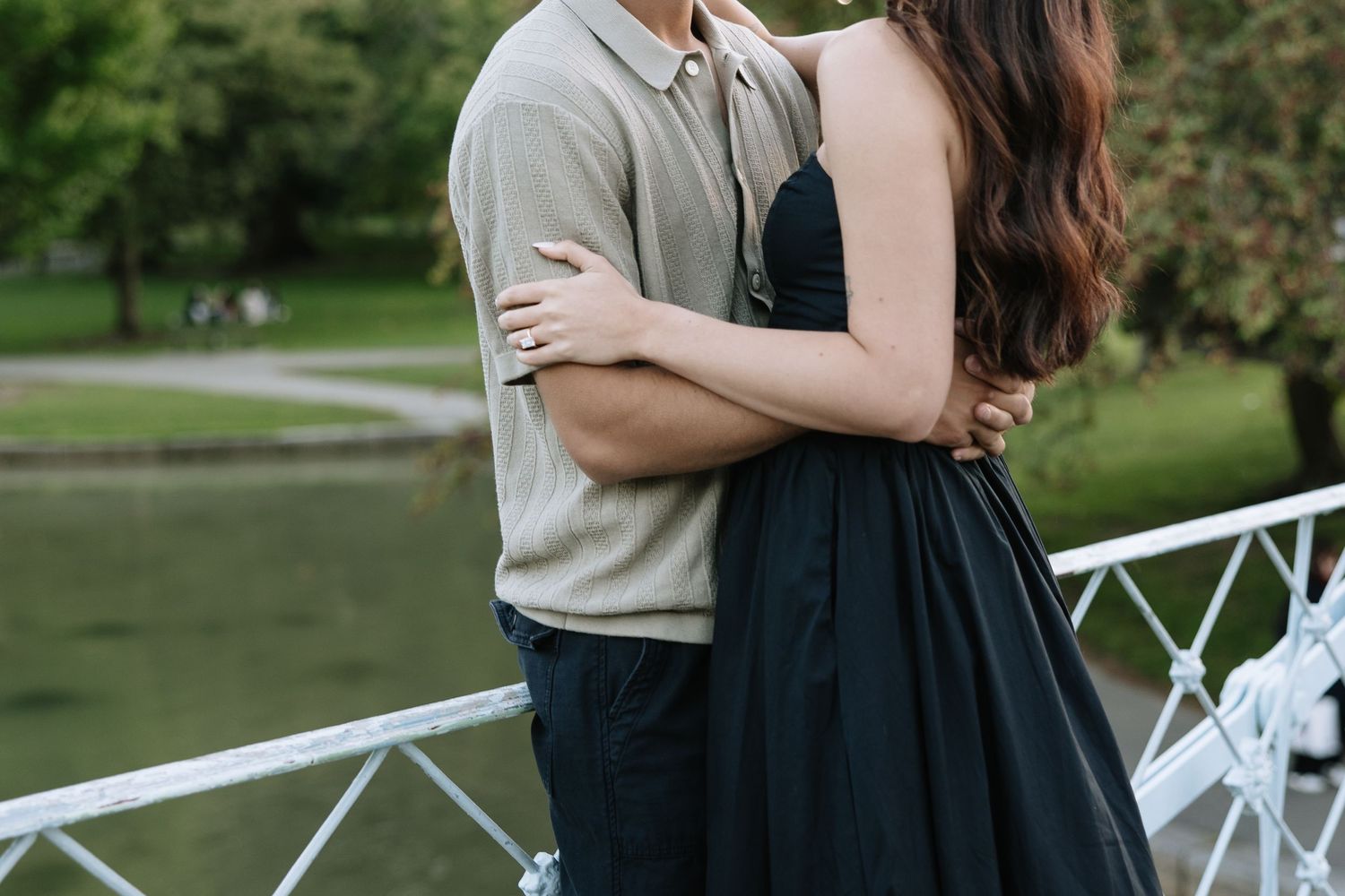 A couple embraces on a bridge in a park with lush greenery in the background.
