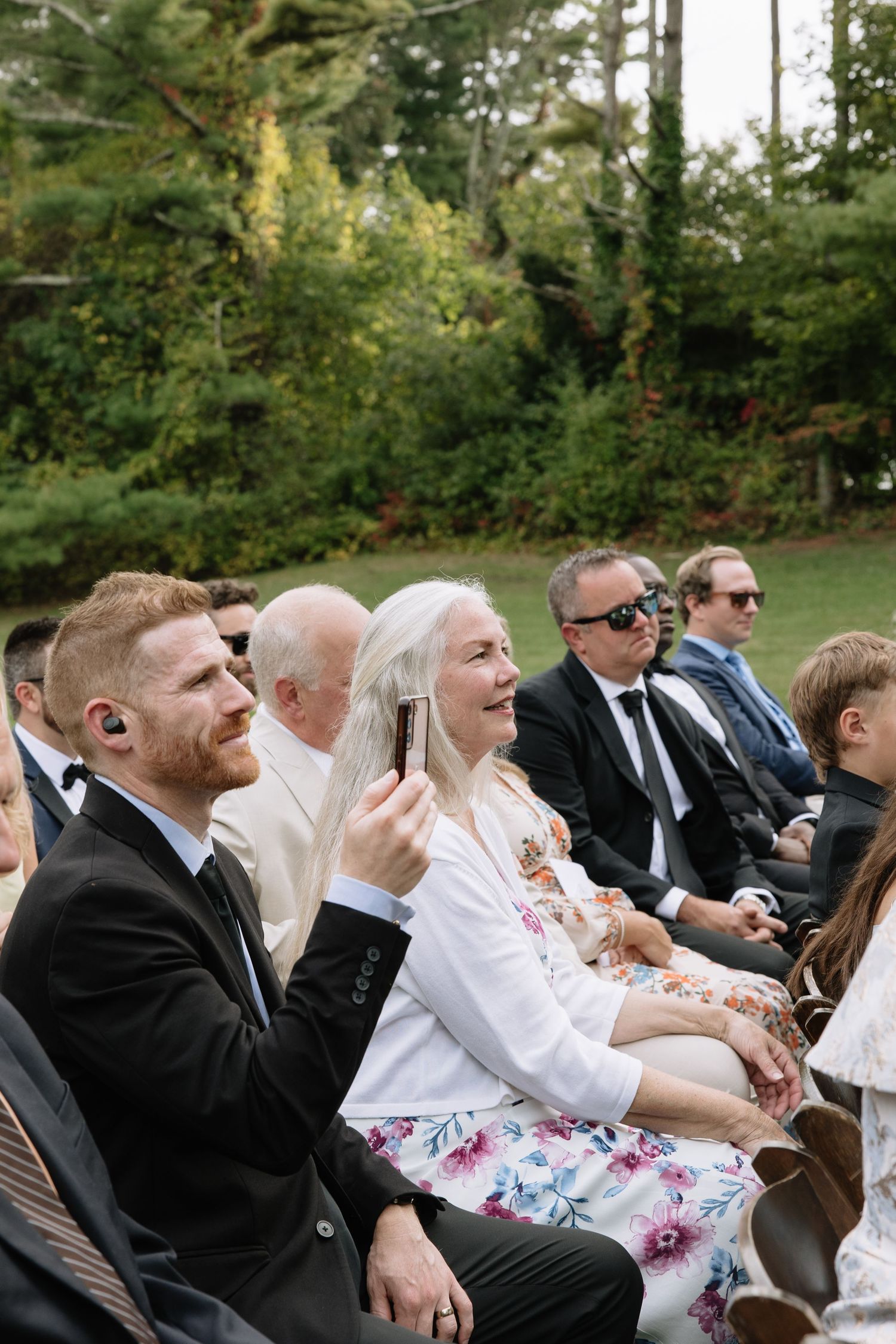 Wedding guests enjoy the outdoor ceremony while seated in garden chairs among pine trees.