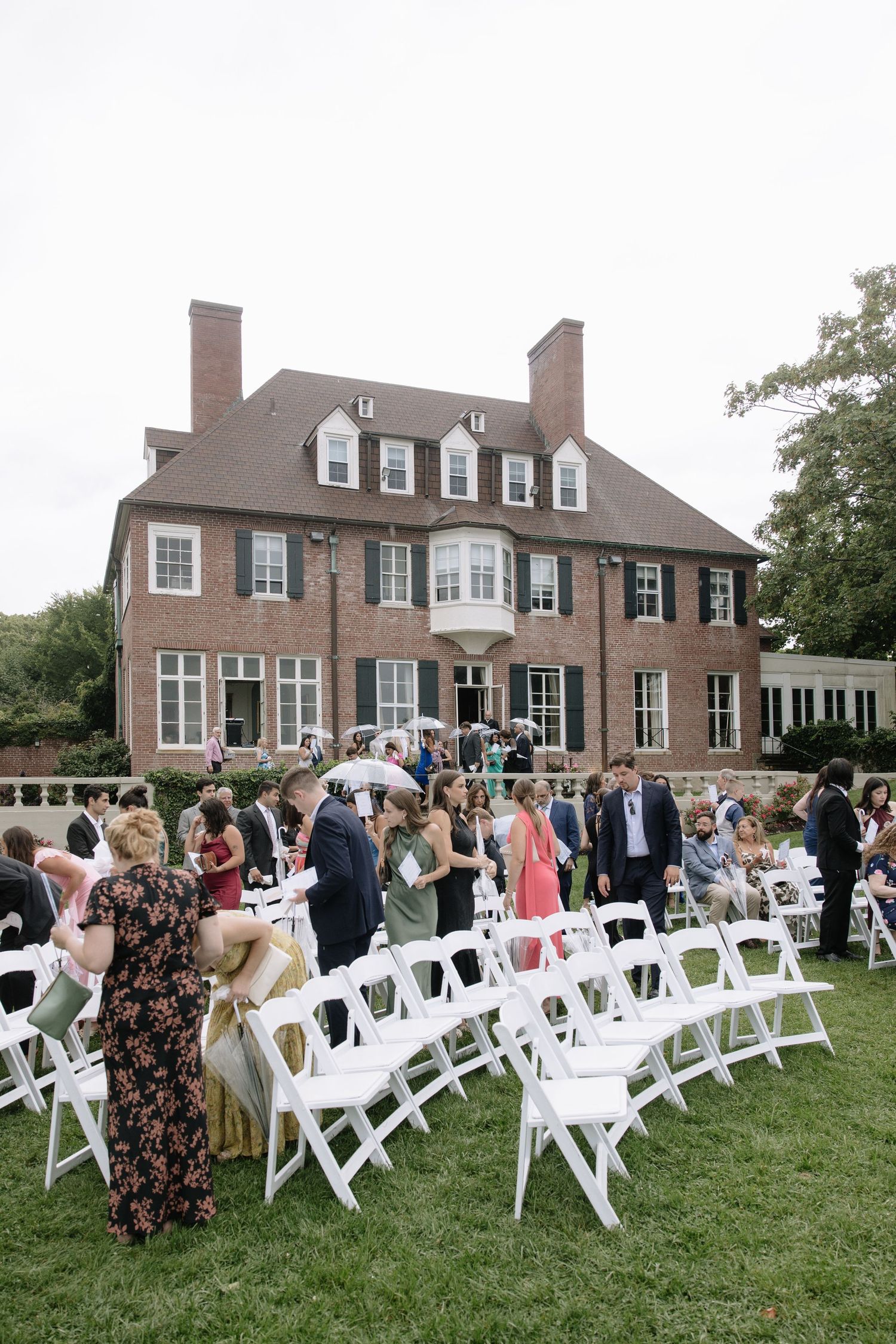 White chairs arranged on a lawn for an outdoor wedding ceremony in front of a large brick mansion.
