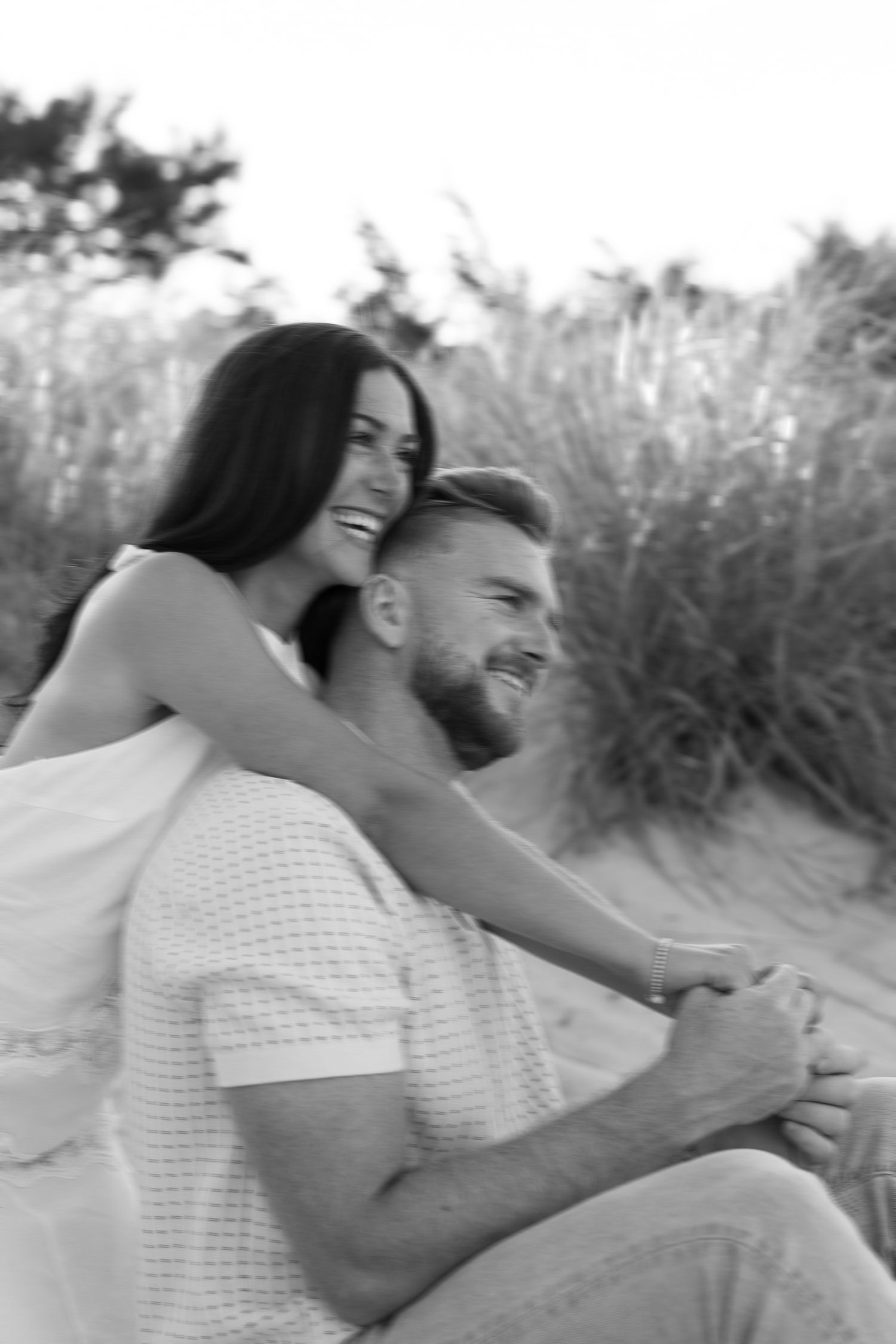 Black and white photo of a smiling couple embracing on sandy beach dunes with tall grass in the background.