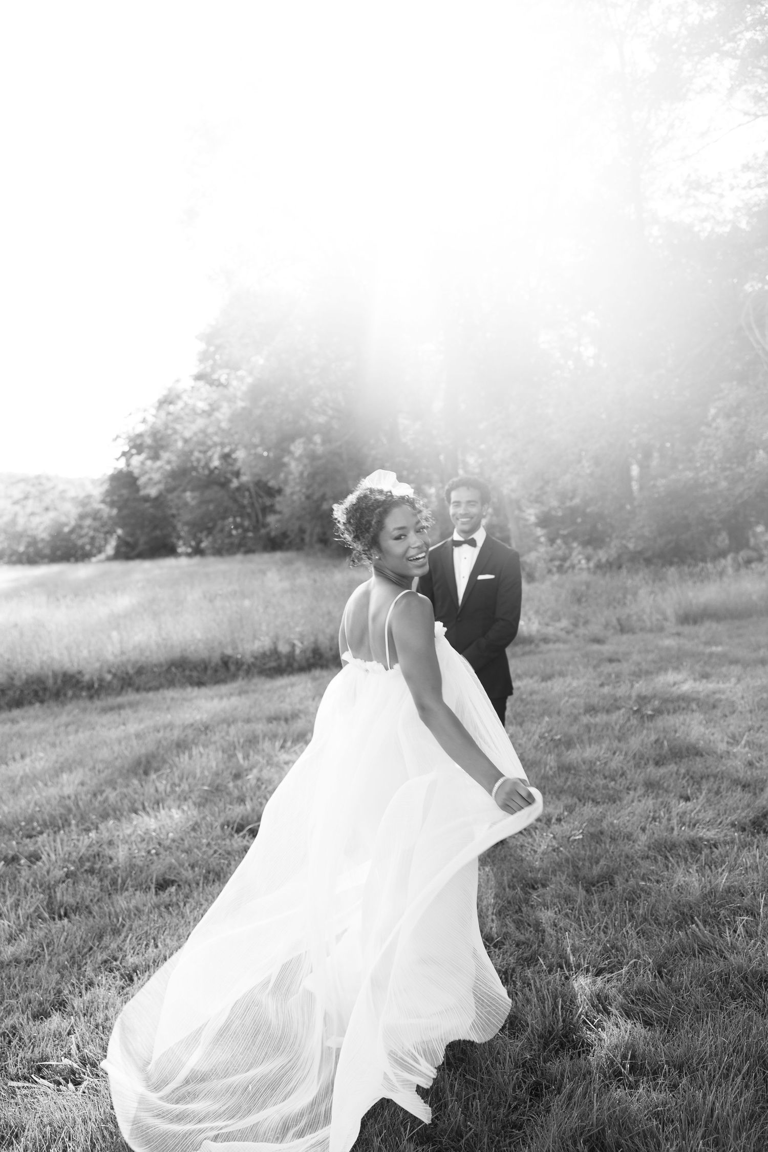 Black and white photo of smiling bride in flowing gown with groom in tuxedo in sunlit field.