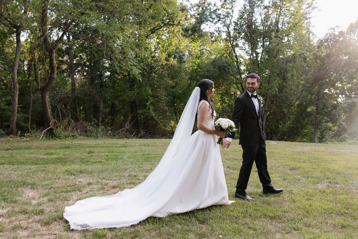 Bride in white gown with long veil walks with groom in tuxedo across green lawn surrounded by lush trees.