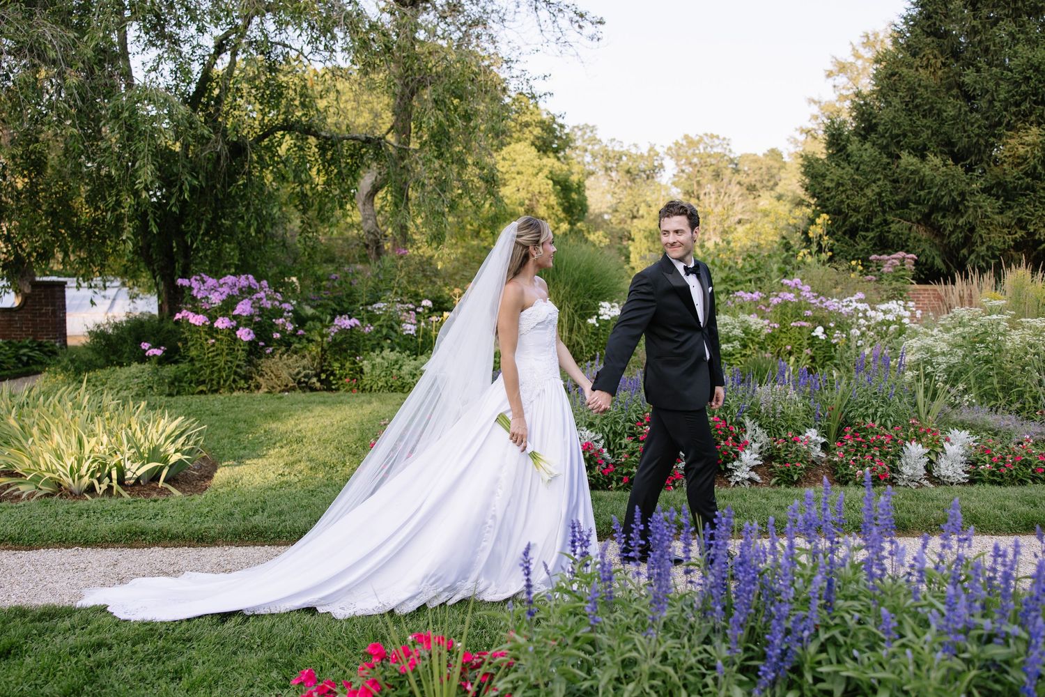 A bride in a white gown and groom in a tuxedo hold hands in a lush garden filled with colorful flowers.
