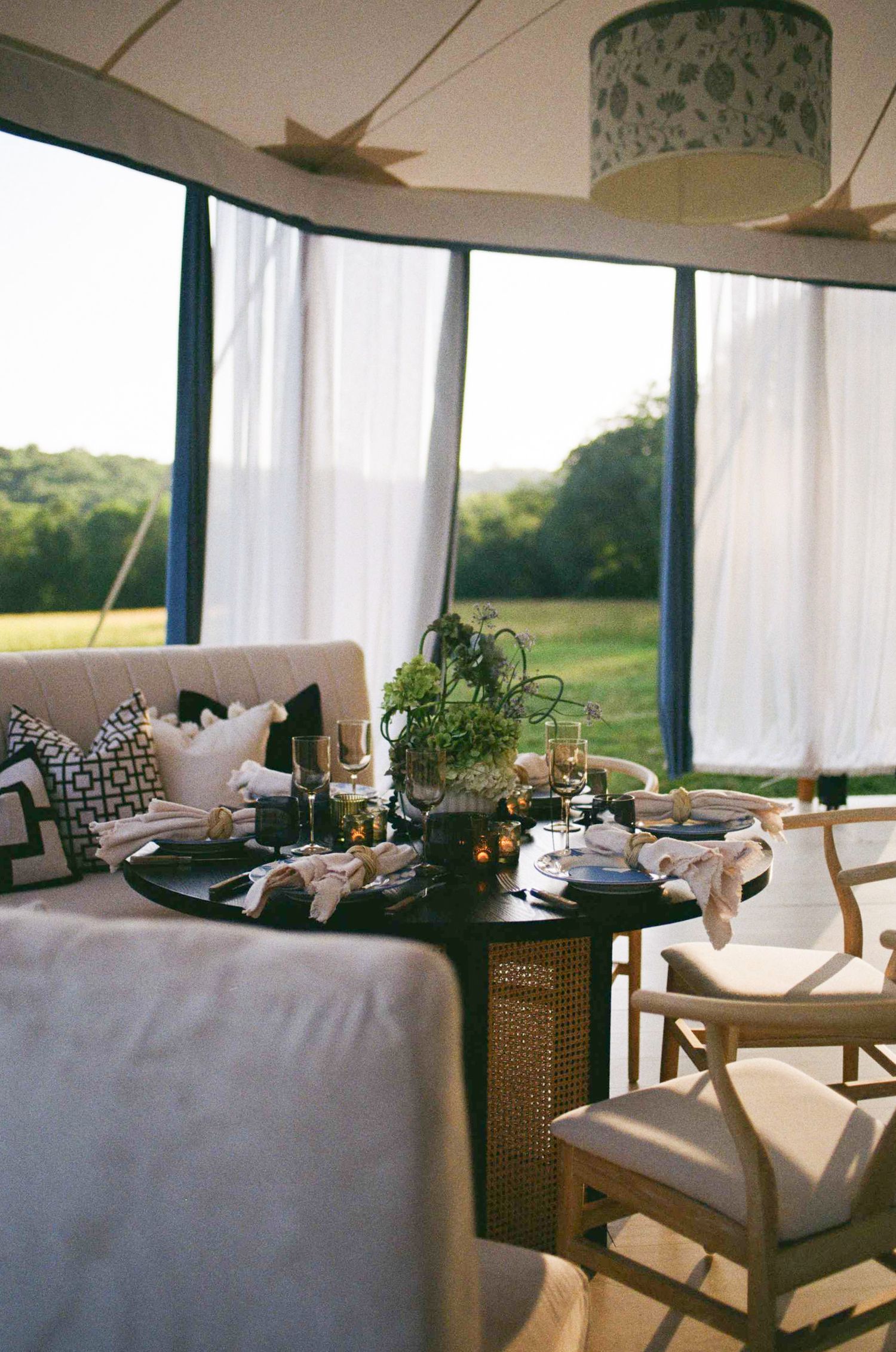 Elegant tent dining setup with dark round table, floral centerpiece, white curtains, and lush green landscape views.