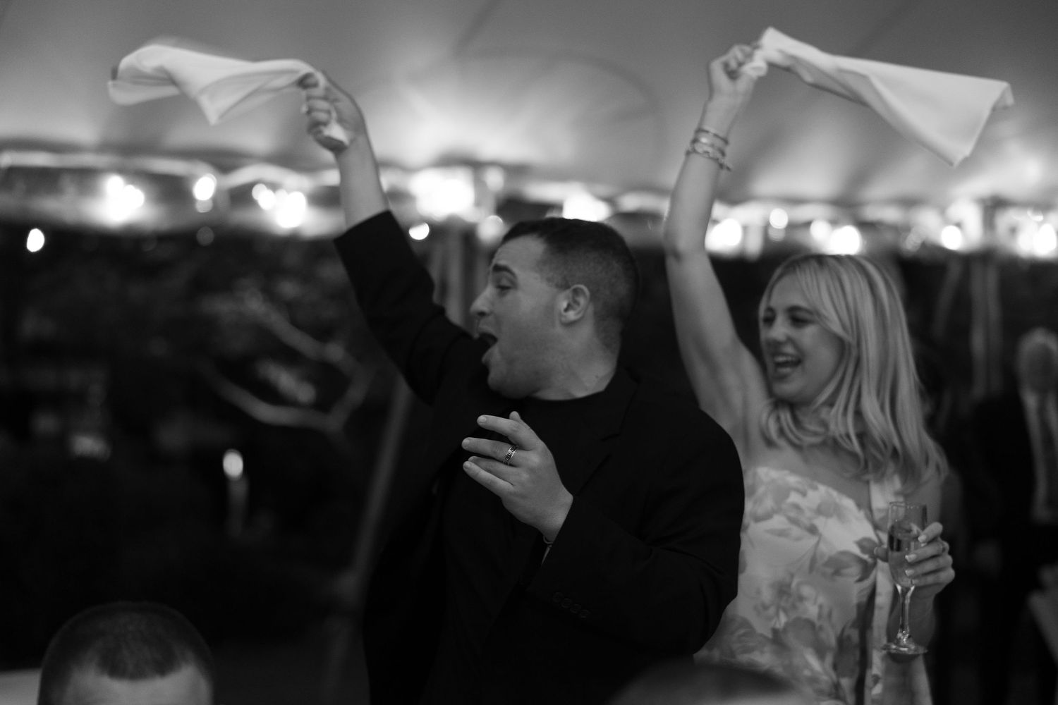 Black and white photo of a joyful couple waving white napkins at a lively wedding reception celebration.