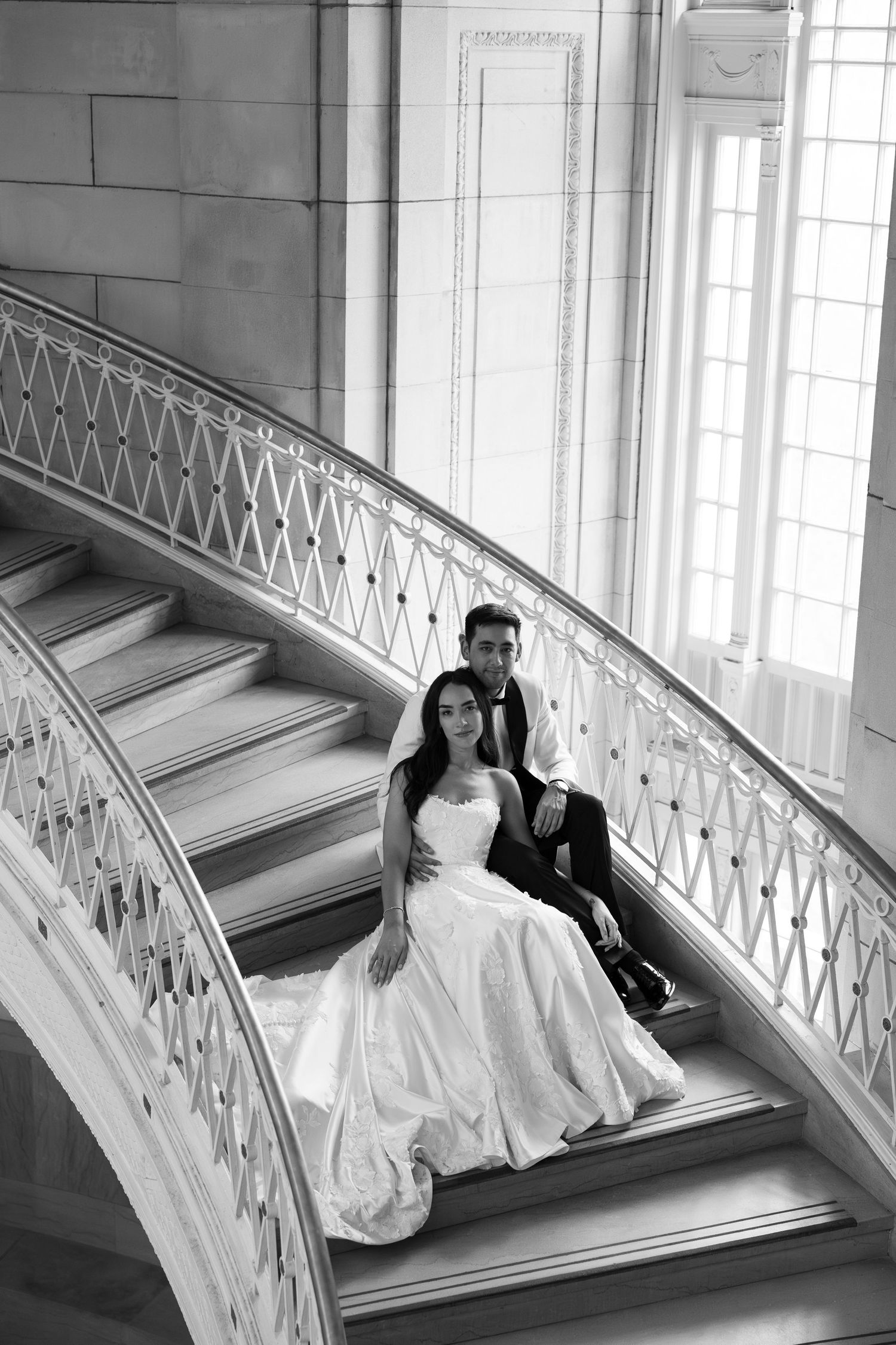 Bride and groom sit elegantly on curved marble staircase in black and white wedding portrait.