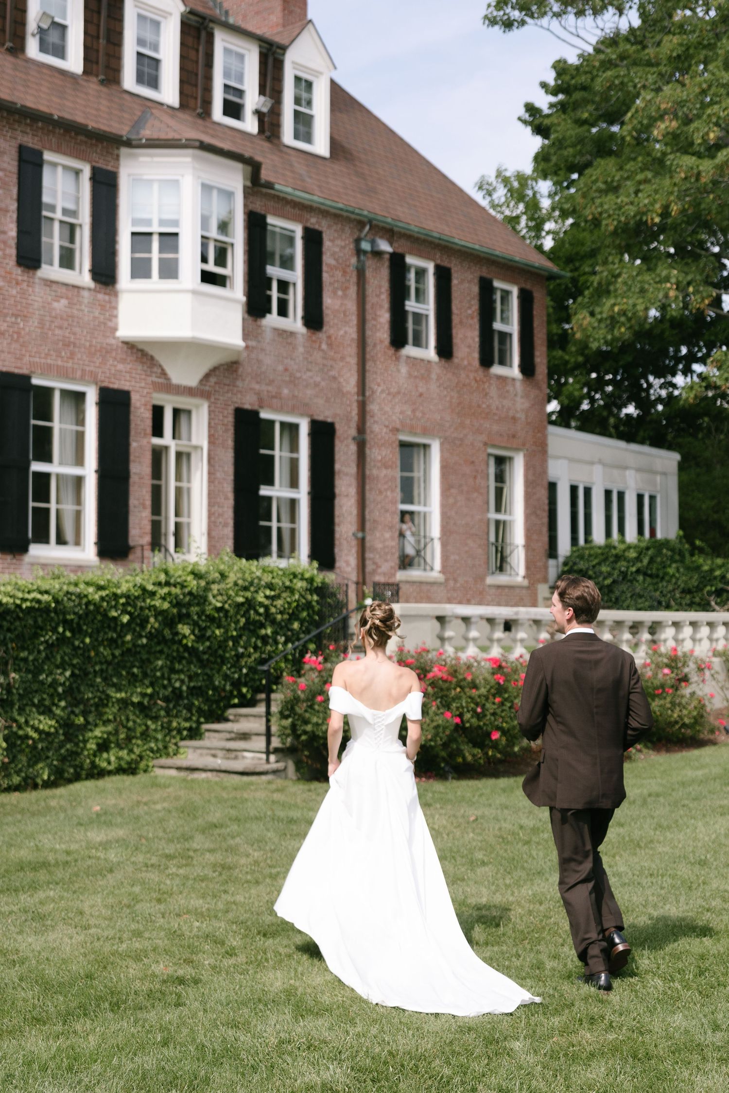 Bride in an off-shoulder white gown and groom in a dark suit walk across a green lawn toward a historic brick estate wedding venue.