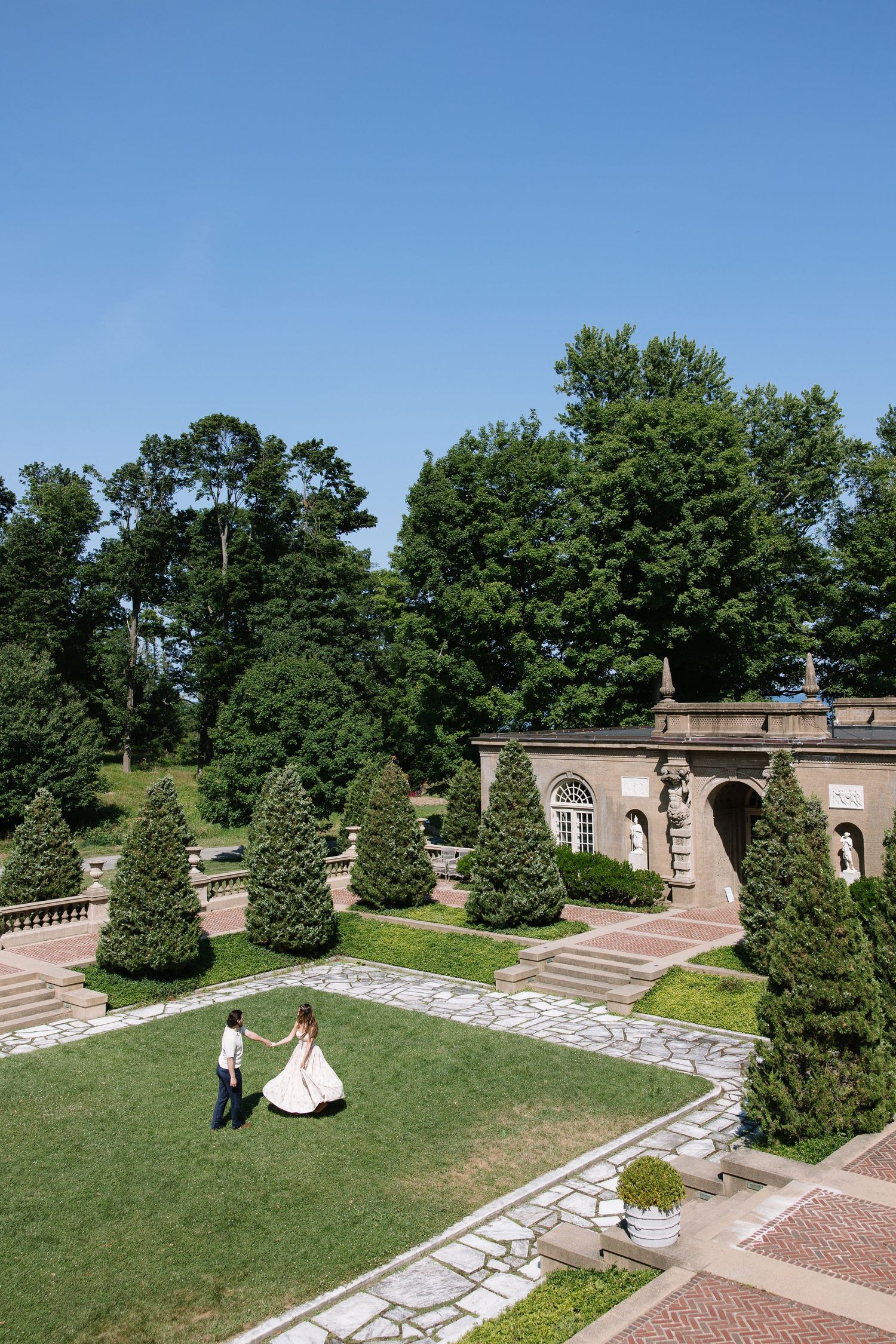 Bride and groom dancing in elegant garden estate with manicured hedges, stone architecture, and lush green trees under blue sky.